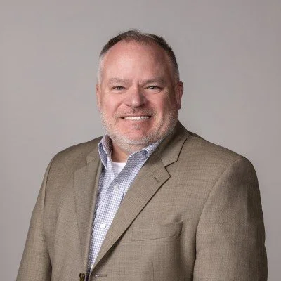 Professional portrait of a smiling middle-aged man with short hair, wearing a beige blazer and a light blue checkered shirt, against a plain gray background.