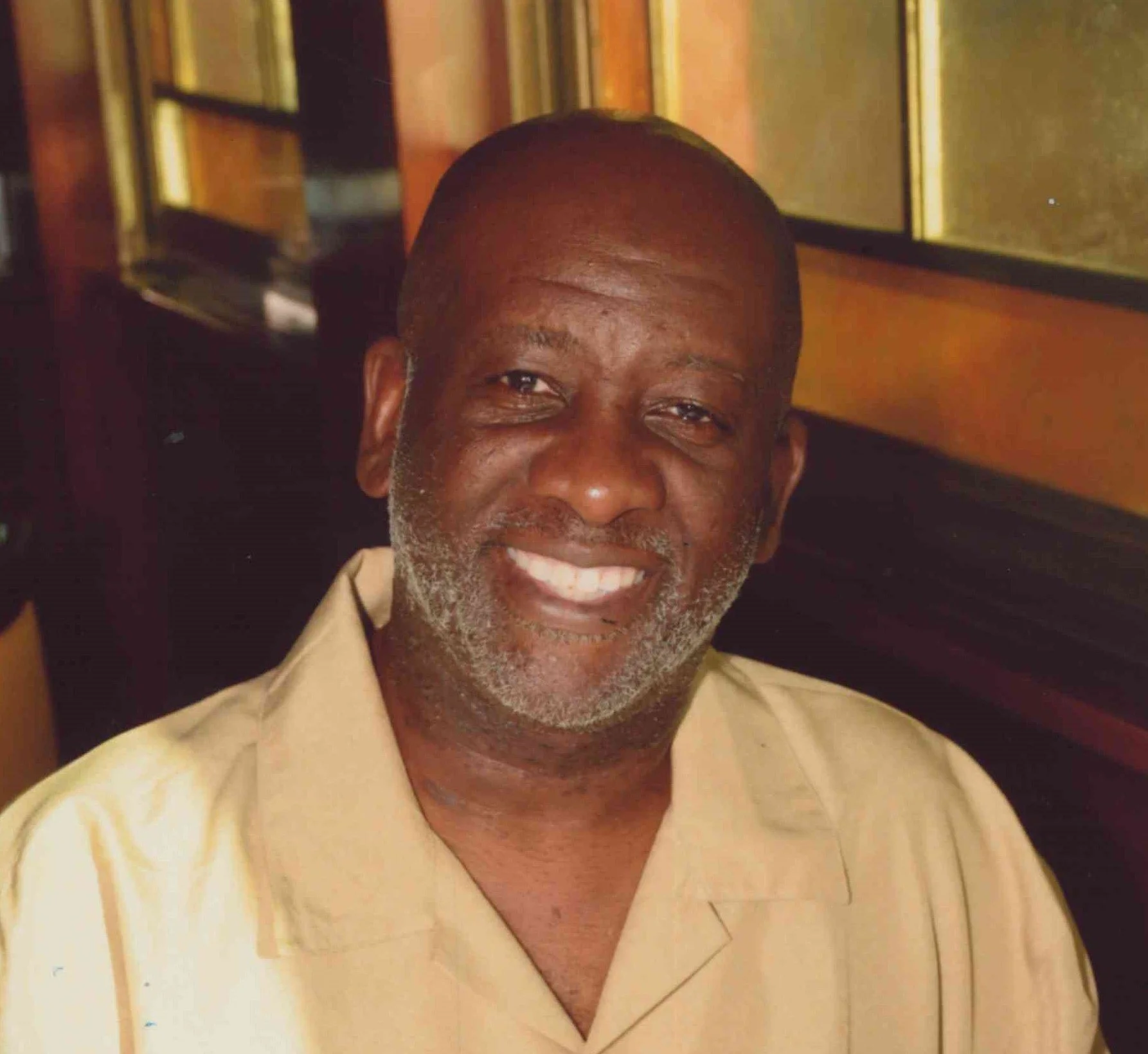 A smiling middle-aged African American man with a beard and bald head, wearing a beige shirt, sitting indoors with a wooden frame and glass behind him.