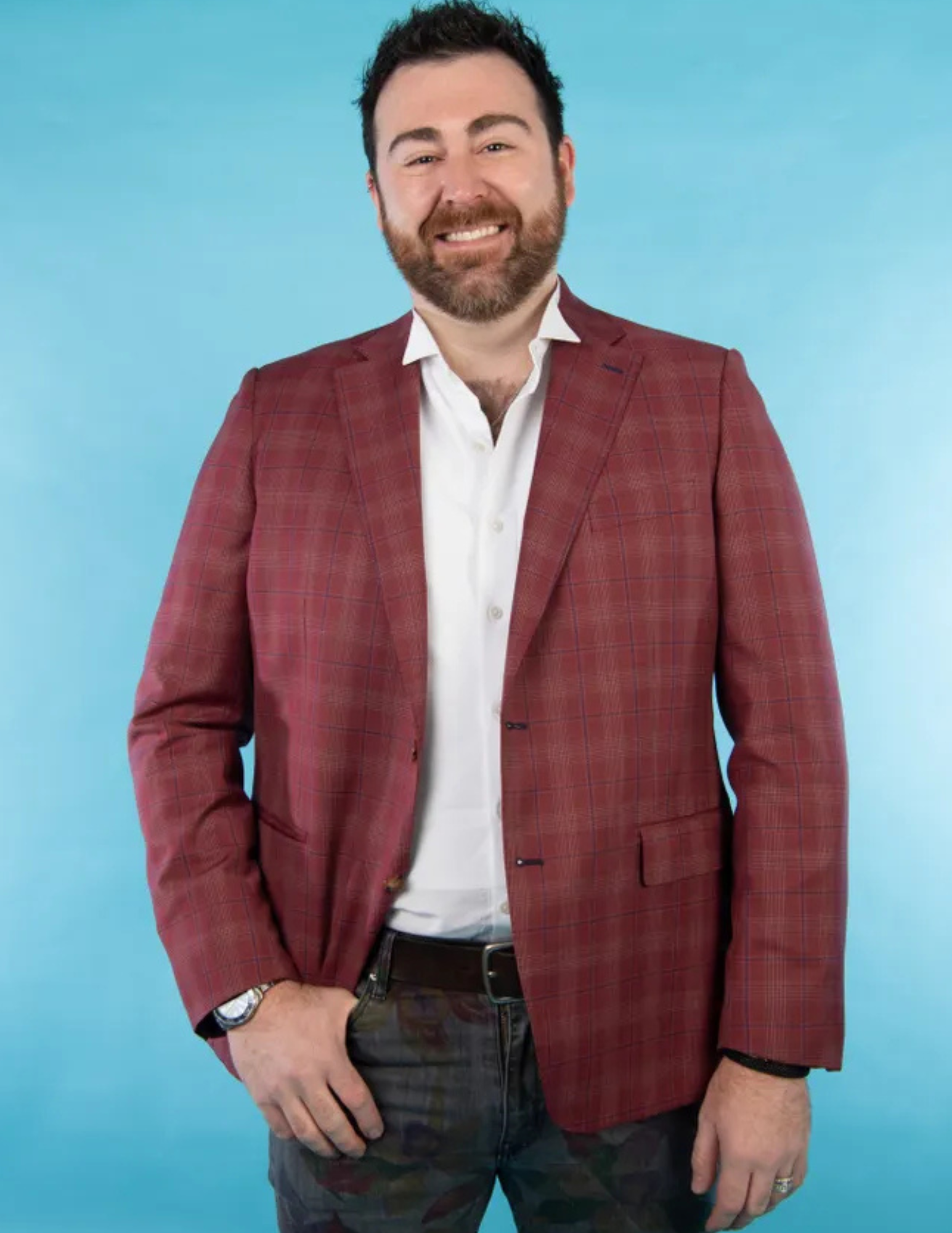 A man with dark hair and a beard, smiling, wearing a red plaid blazer, white dress shirt, and dark pants, standing against a light blue background.