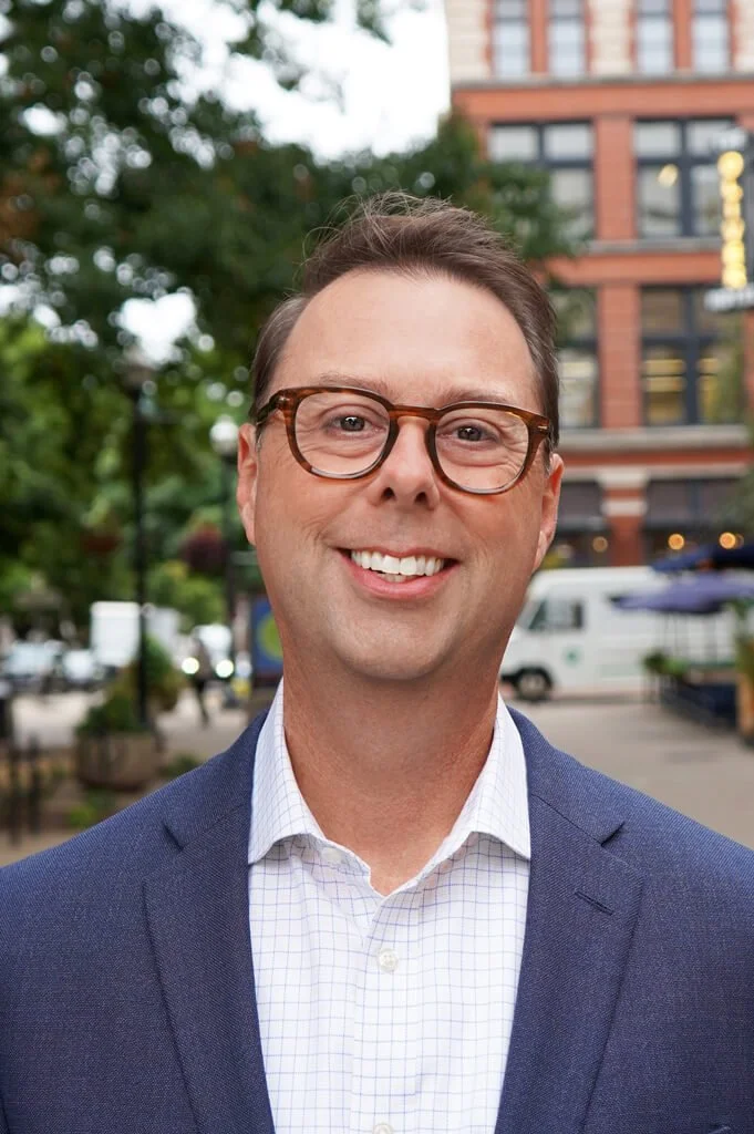 Man in glasses smiling outdoors in an urban area with trees and buildings in the background.