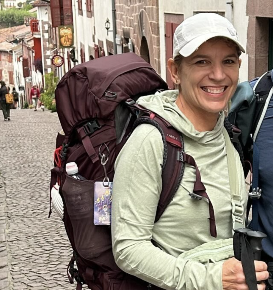 Smiling woman with a backpack, wearing a light-colored hoodie and cap, on a cobblestone street in a European town.