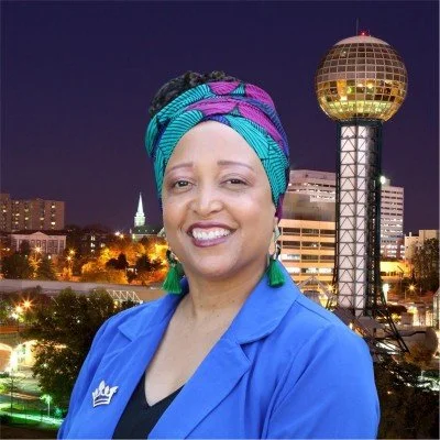 A woman smiling in front of a city skyline with a distinctive tall, spherical building at night. She is wearing a colorful headwrap, green earrings, and a blue jacket.