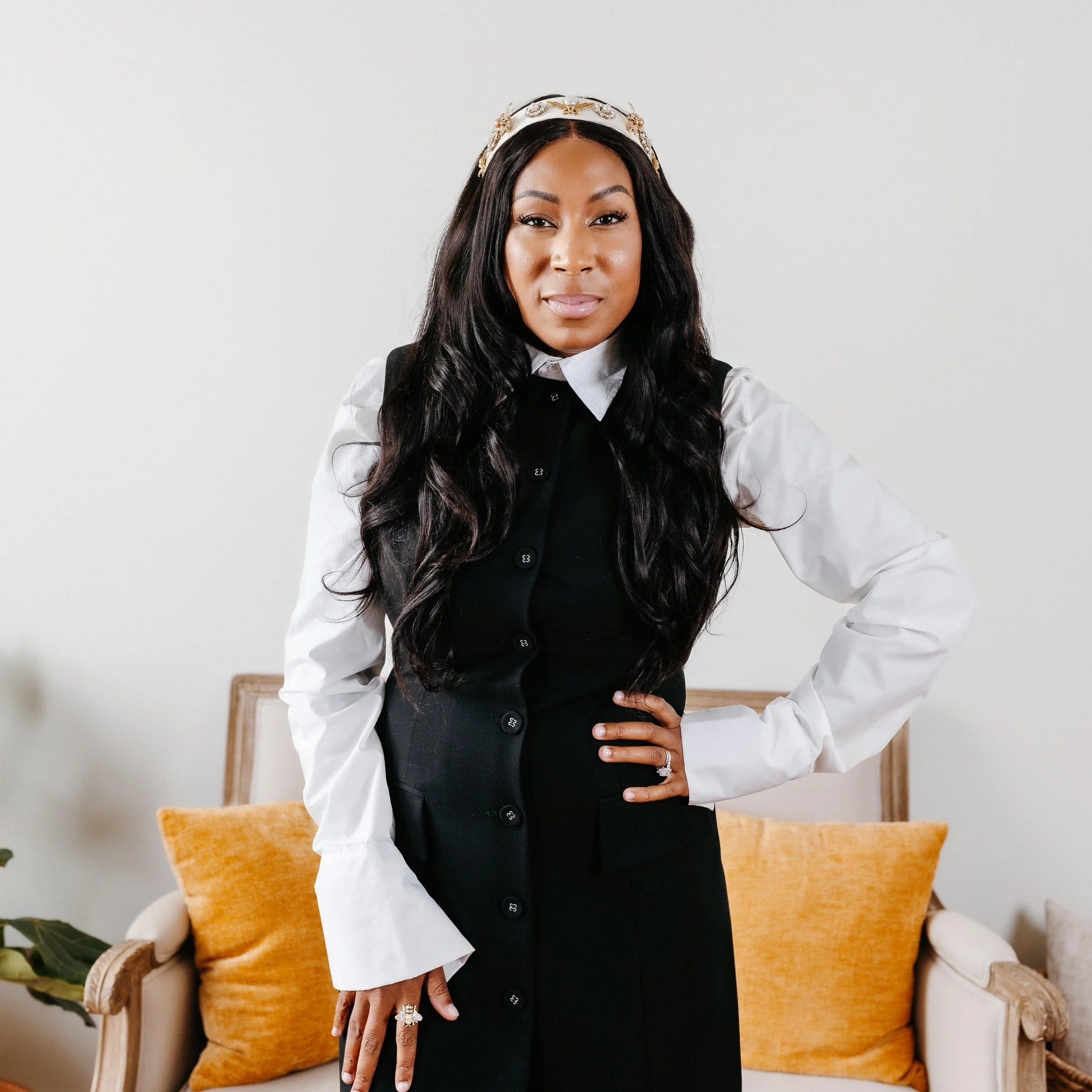 A woman with long, wavy black hair, wearing a white blouse and black vest, standing confidently in front of a beige sofa with orange pillows, posing with one hand on her hip.