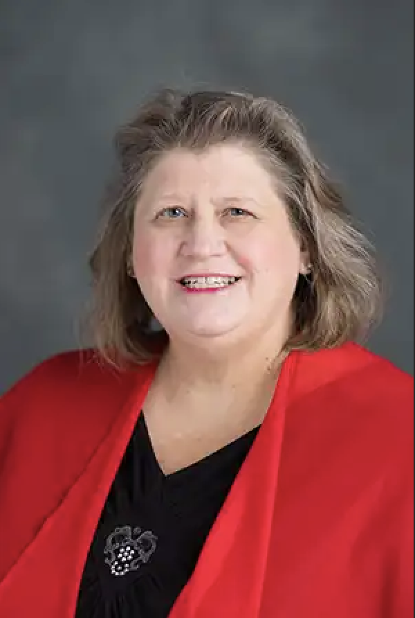 A smiling woman with shoulder-length gray hair, wearing a red jacket over a black top, against a gray background.