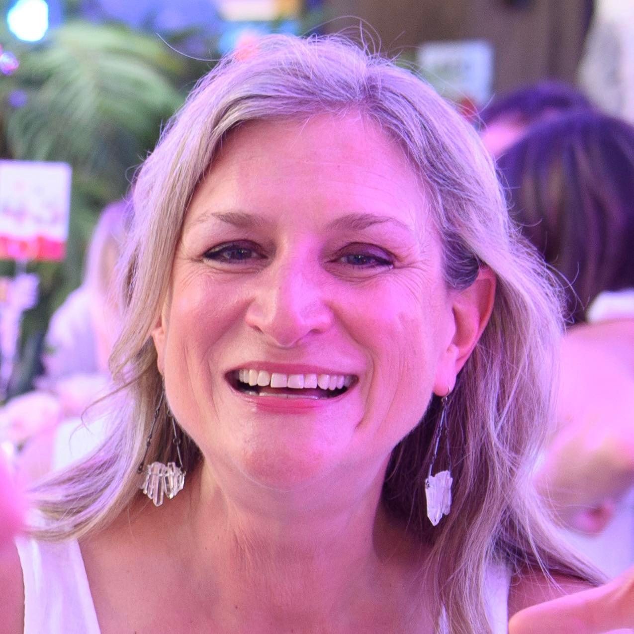 A smiling woman with shoulder-length blonde hair, wearing earrings, in a setting with colorful lighting and blurred background.