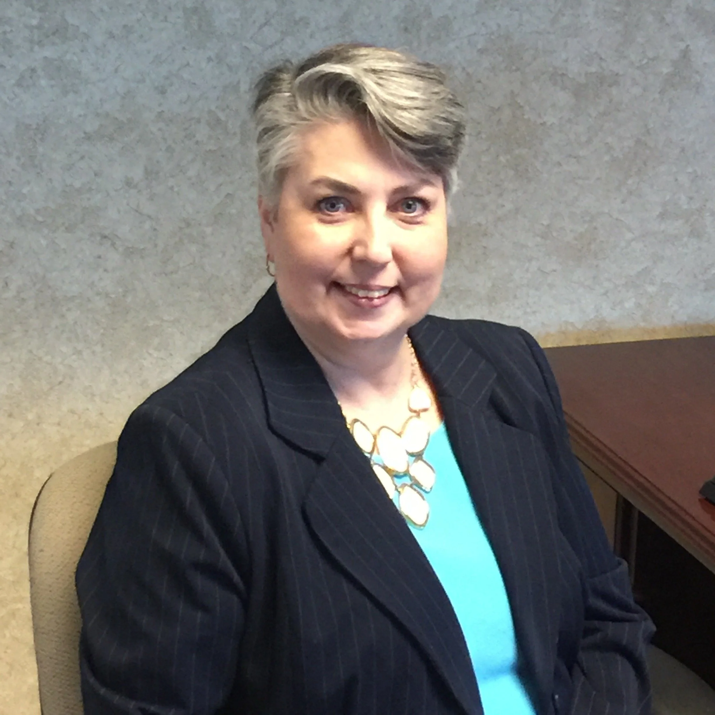 A smiling woman with short blonde hair, wearing a dark pinstripe blazer, a light blue top, and a large necklace, seated at a desk against a textured gray wall.