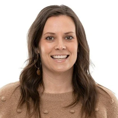 A woman with long wavy brown hair, wearing a tan sweater, smiling against a white background.