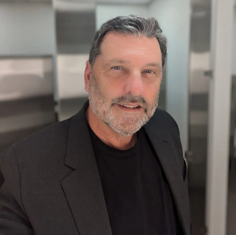 A middle-aged man with gray hair and beard, wearing a black shirt and blazer, smiling indoors.