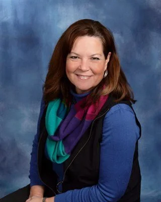 A woman with long brown hair, wearing a blue shirt, a colorful scarf, and earrings, smiling at the camera against a blue background.
