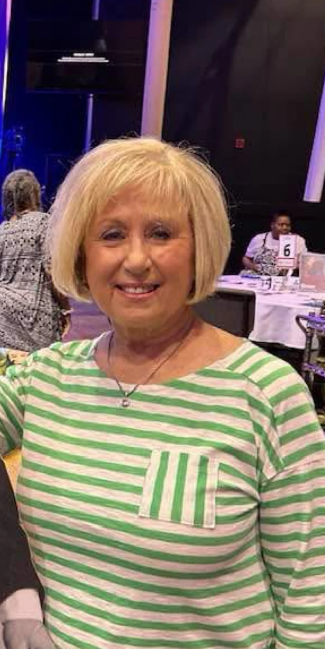 A smiling woman with blonde hair in a short bob, wearing a green and white striped shirt with a front pocket, and a necklace. She is indoors at a social event with tables and people in the background.