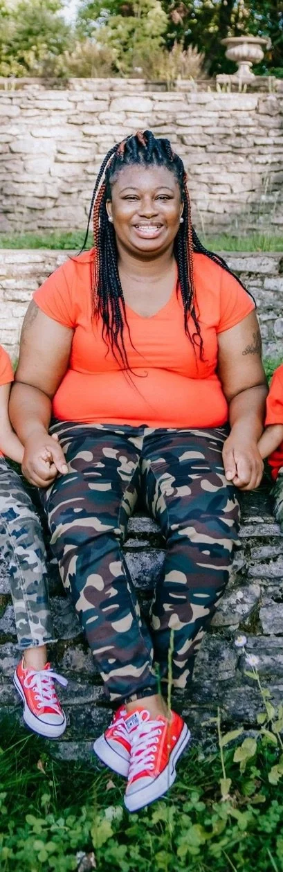 A woman sitting on a stone bench outdoors, wearing an orange shirt, camouflage pants, and red sneakers, smiling at the camera.