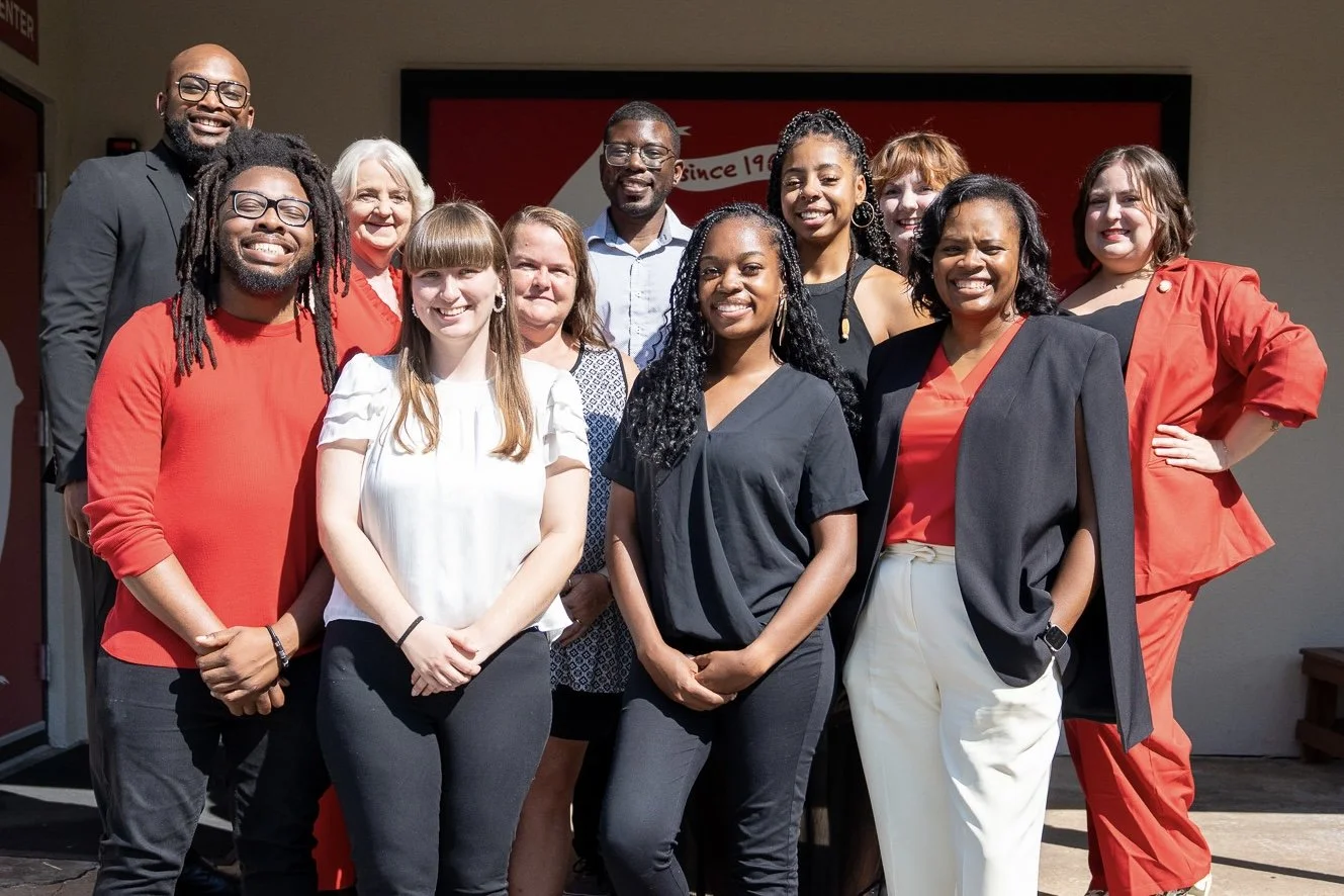 A group of diverse smiling people, including men and women, standing outdoors in front of a wall with a red and white banner. They are dressed in business casual and formal clothing.