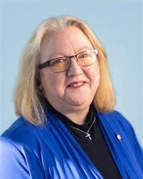 Headshot of a professional smiling White woman with shoulder-length blonde hair, wearing glasses, a dark top with a green jacket, also wearing a necklace with a cross, against a light green background.