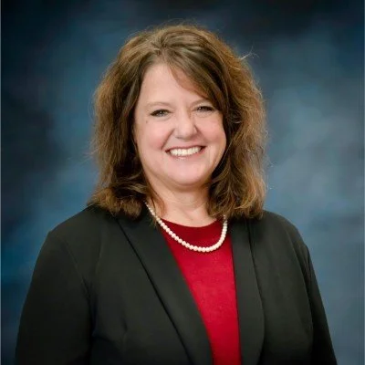 A professional White woman with shoulder-length brown wavy hair, smiling, wearing a red sweater, a blue jacket and a white pearl necklace, against a lighter blue background.