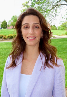 A professional White woman who is smiling, with long wavy-brown hair, wearing a white top and a light beige jacket, against an outdoors backdrop.