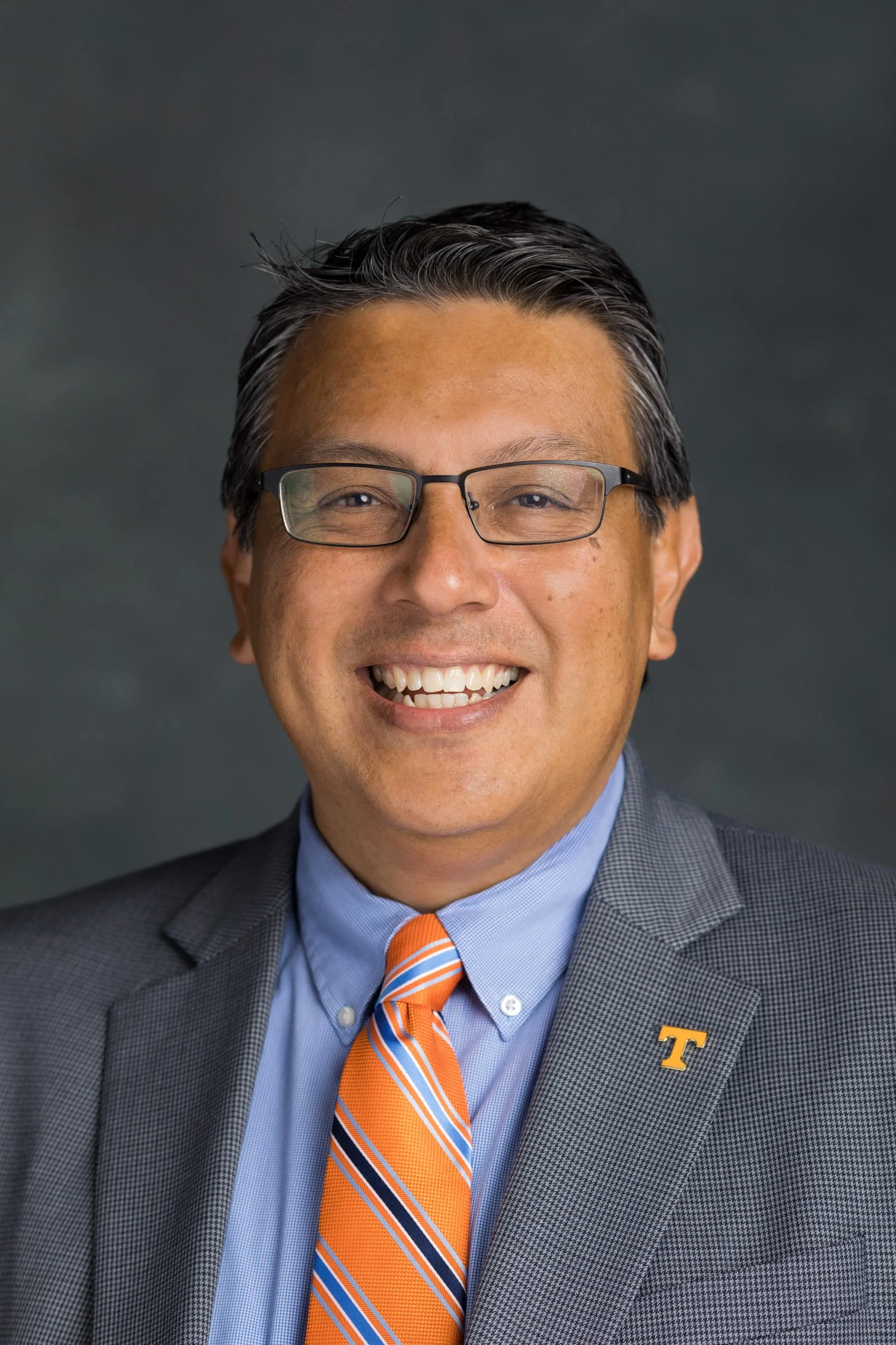 A professional Hispanic man in glasses, smiling, wearing a light blue shirt, a light blue blazer and a colorful pink tie with blue stripes, set off with a University of Tennessee lapel pin.