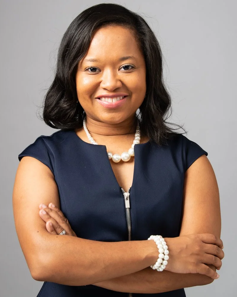 A professional Black woman, smiling, with shoulder-length dark hair, adorned with a pearl necklace and a pearl bracelet. against a light grey background.