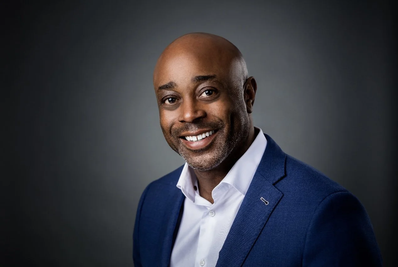 Headshot of a professional Black man with a broad smile, a shaved head, wearing a white business shirt and a blue blazer, against a warm background.