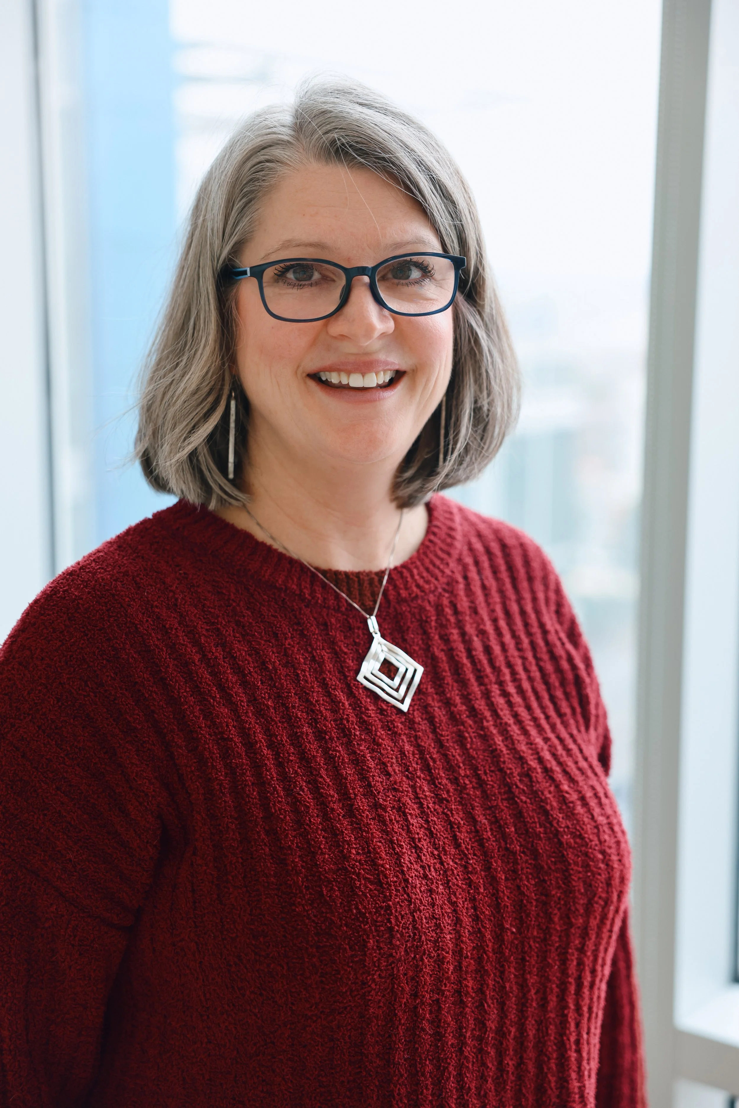 A professional smiling White woman with shoulder-length grey hair, wearing glasses, a red sweater, a necklace with a lovely pendant, earrings, in a setting with colorful lighting and blurred background.