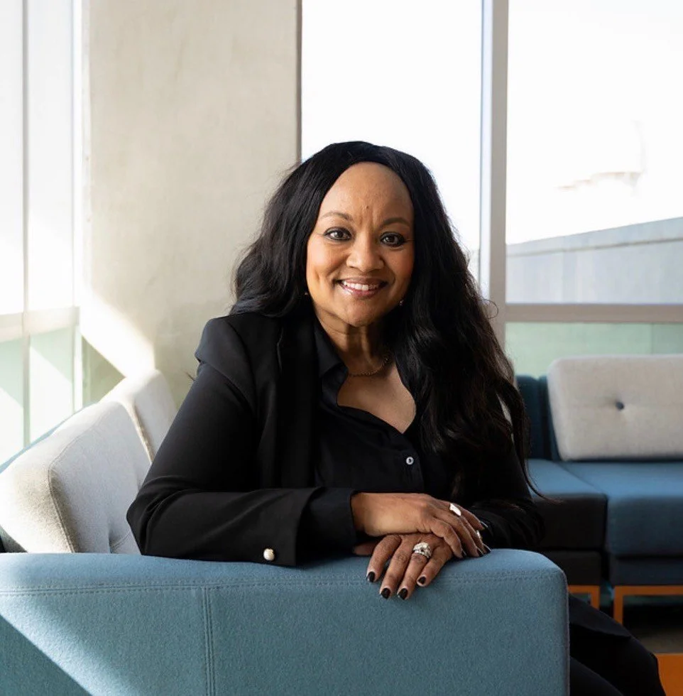 A professional Black woman with long, wavy black hair, smiling, wearing a dark blue blouse and jacket, seated in a teal colored chair, with a window and white wall in the background.