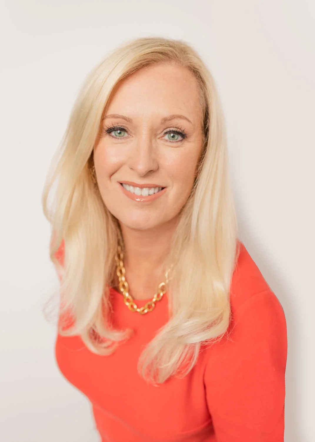 A smiling professional White woman with long blonde hair, wearing a peach sweater and a lovely gold necklace, against a white background.