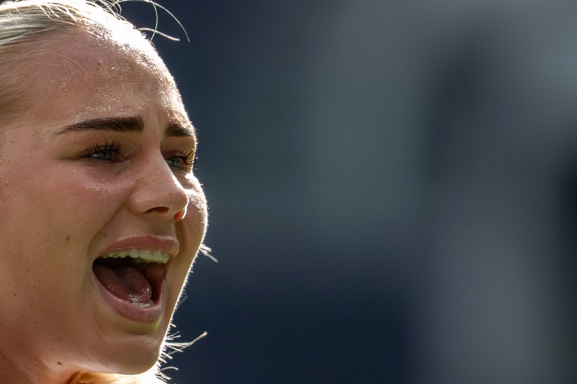 Close-up of a woman with blonde hair, smiling and with her mouth open, showing her teeth and tongue. She has makeup on and is in an outdoor setting with a dark background.