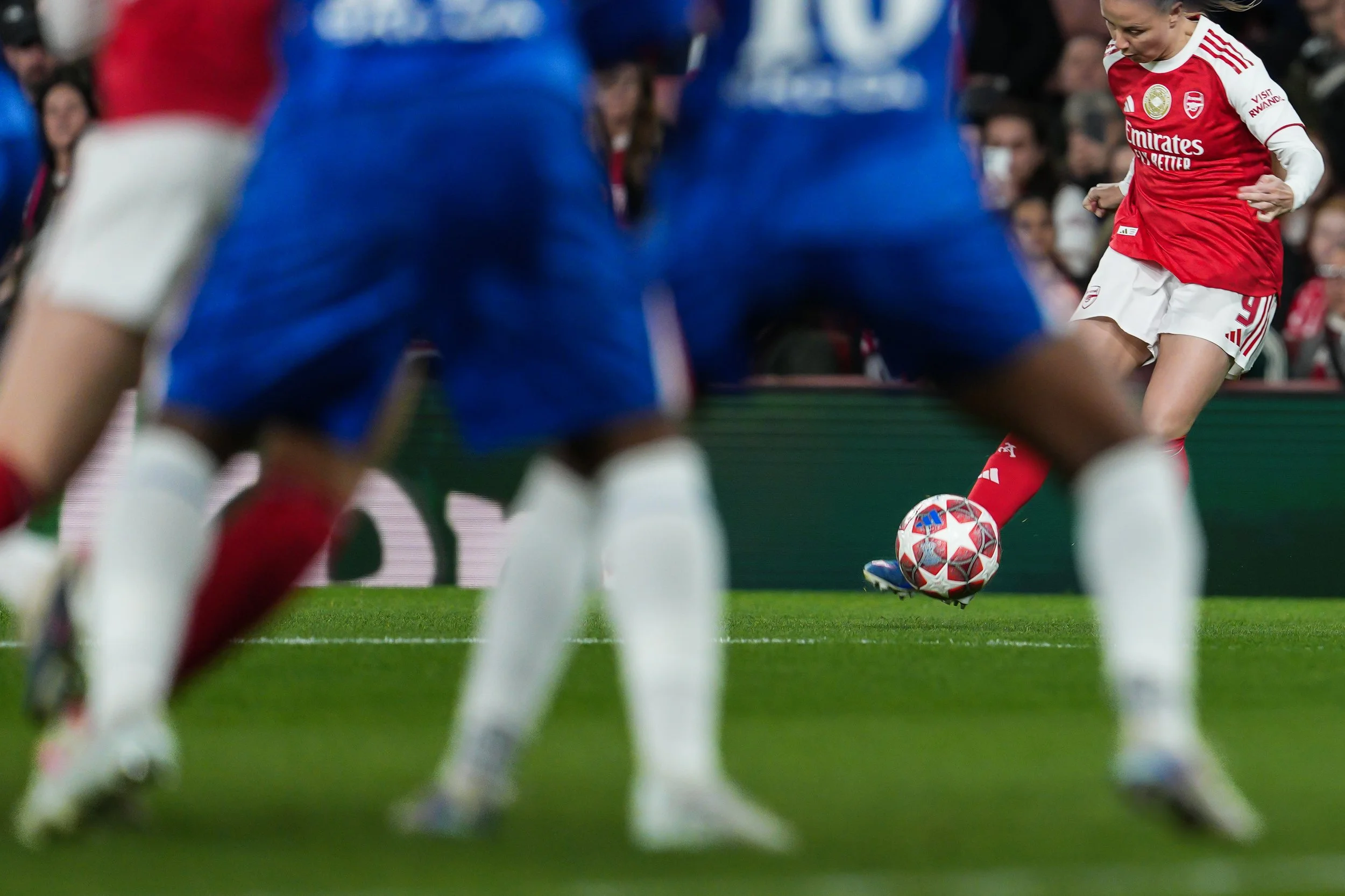A soccer game with players in red and white jerseys, focusing on a player in red kicking a ball, viewed from behind players in blue jerseys.