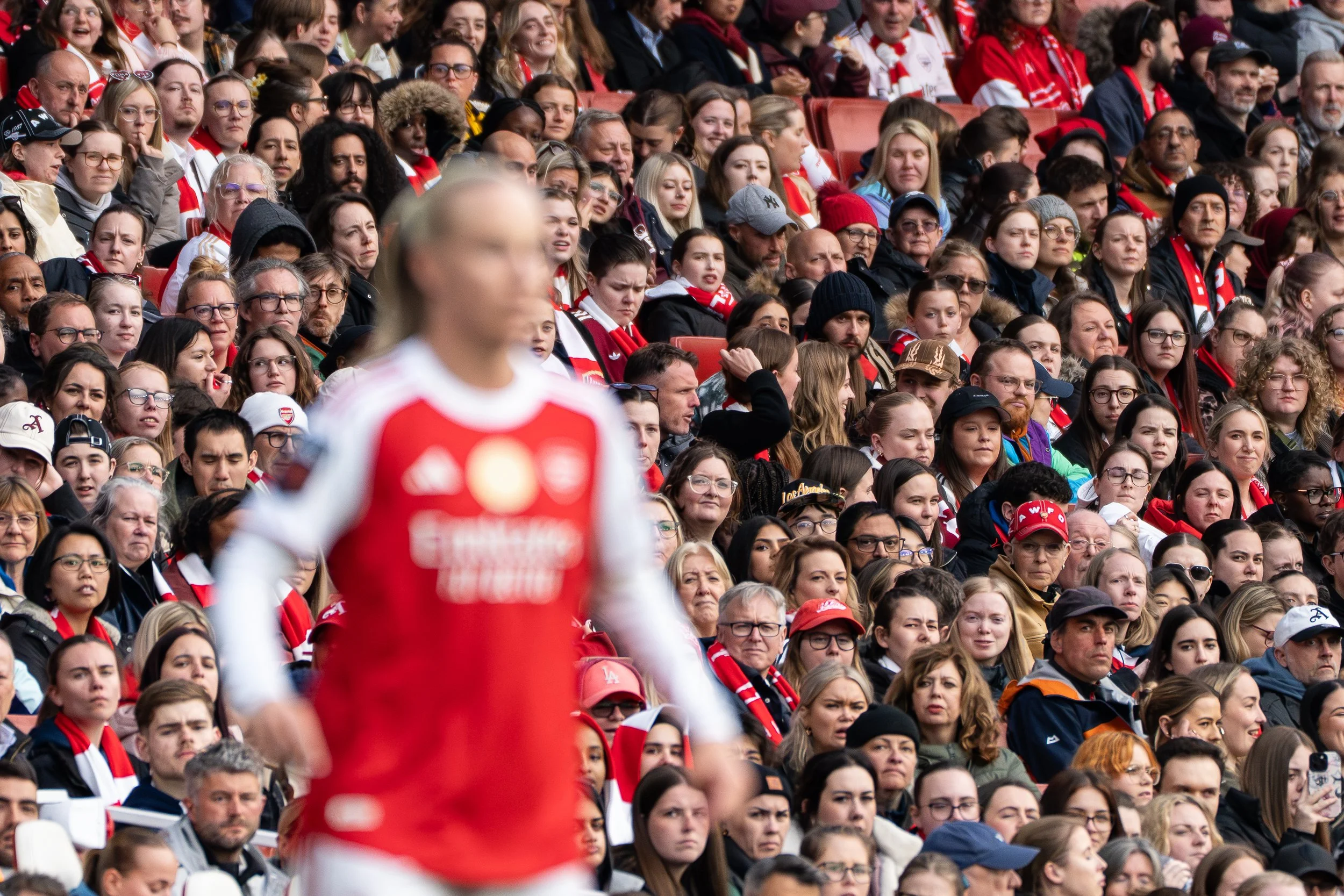 A large crowd of people seated outdoors, many wearing red and white clothing and scarves, watching an event. In the foreground, a blurred person in a red and white shirt is visible, possibly a player or participant.