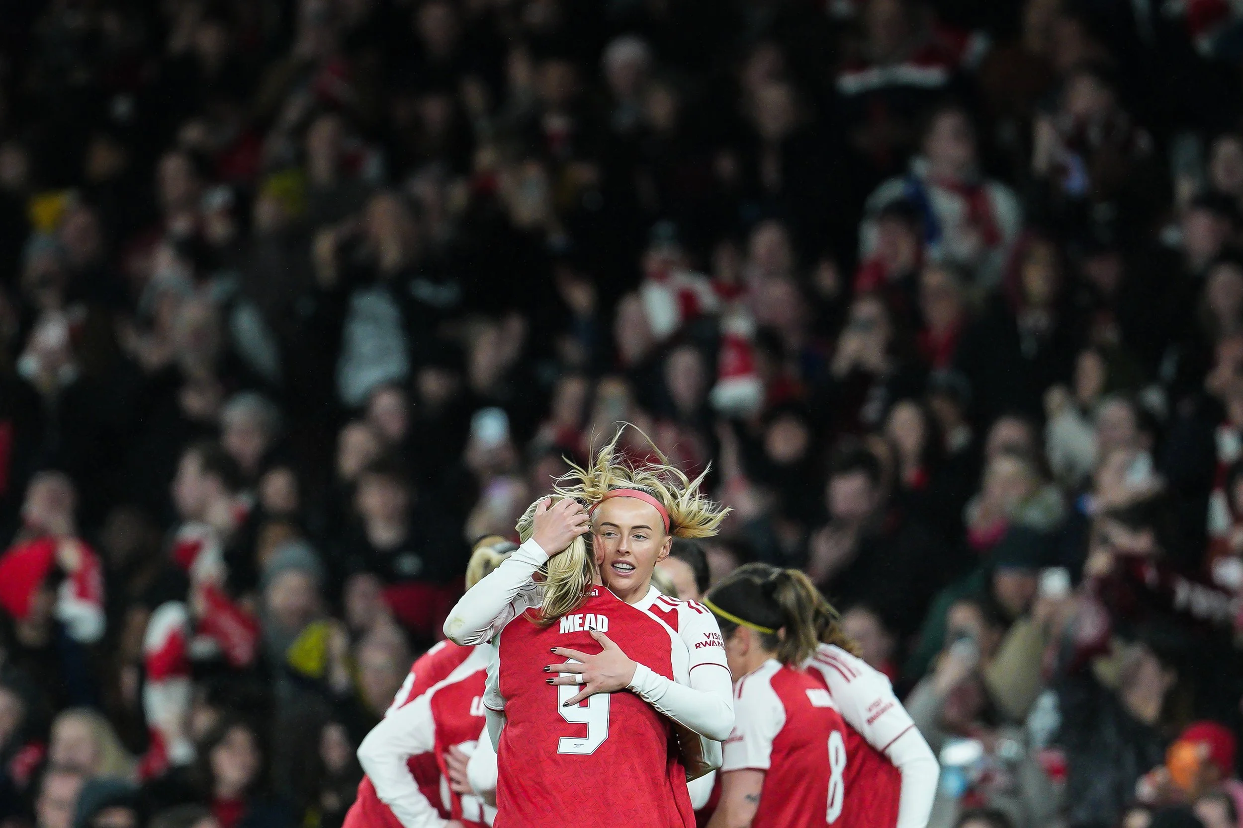 A soccer player with long blond hair, wearing a red and white jersey, is celebrating among teammates on the field at a crowded stadium.