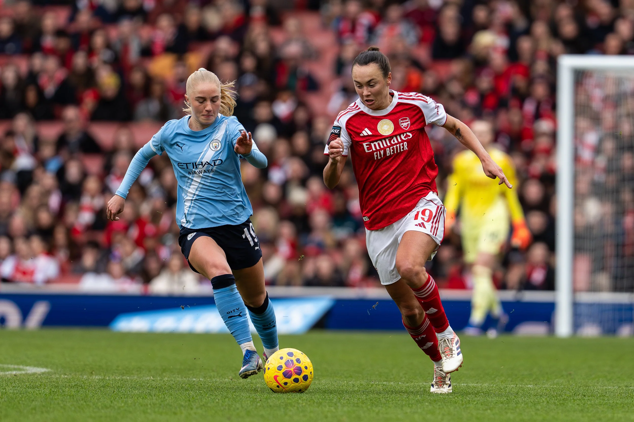 Two female soccer players competing for the ball during a match. One player is in a light blue jersey and the other in a red and white jersey. They are on a green field with a blurred crowd in the background.