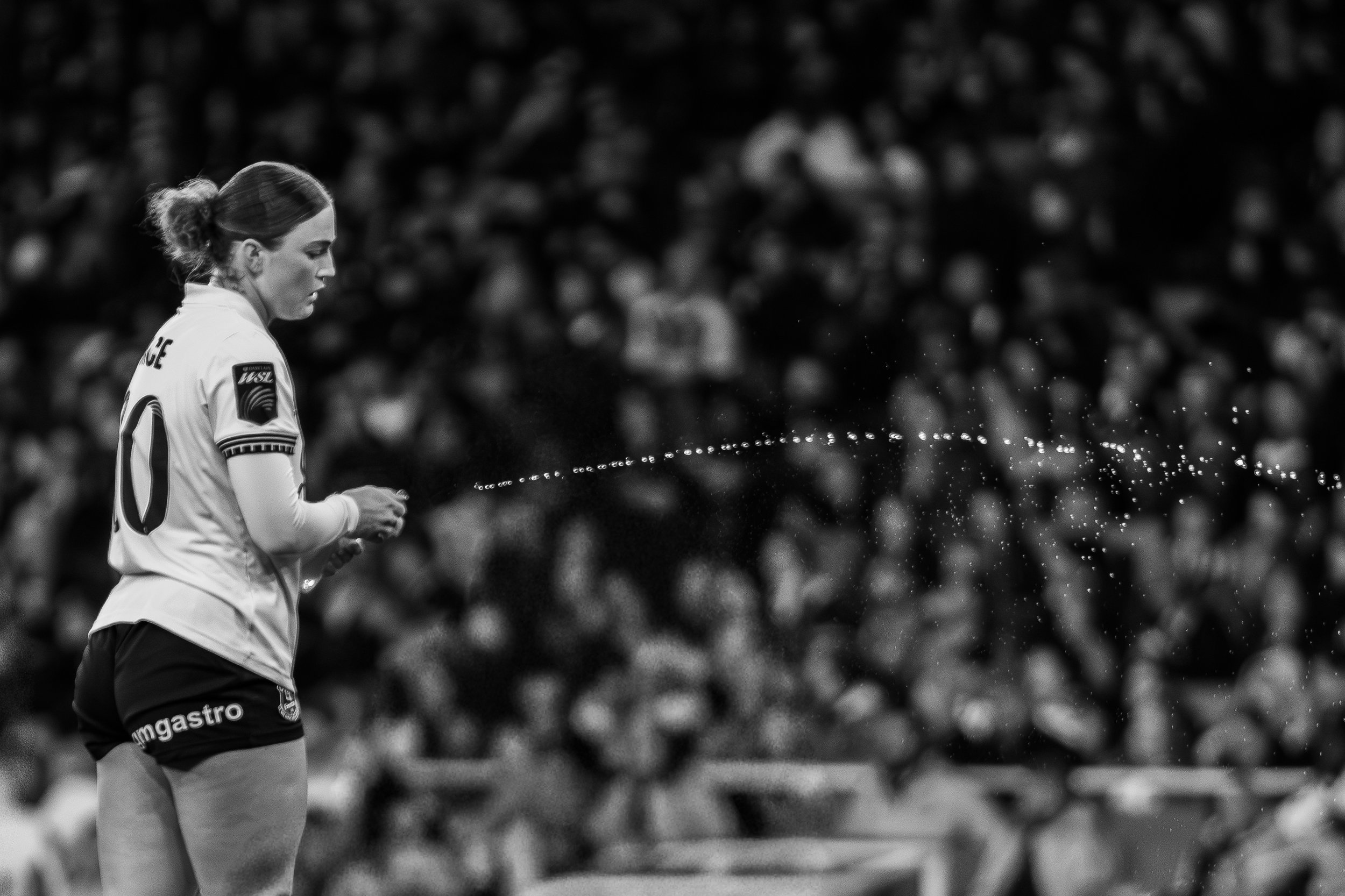 A female soccer player in a jersey standing on the field, with spectators blurred in the background, spraying water or spray from her hand.