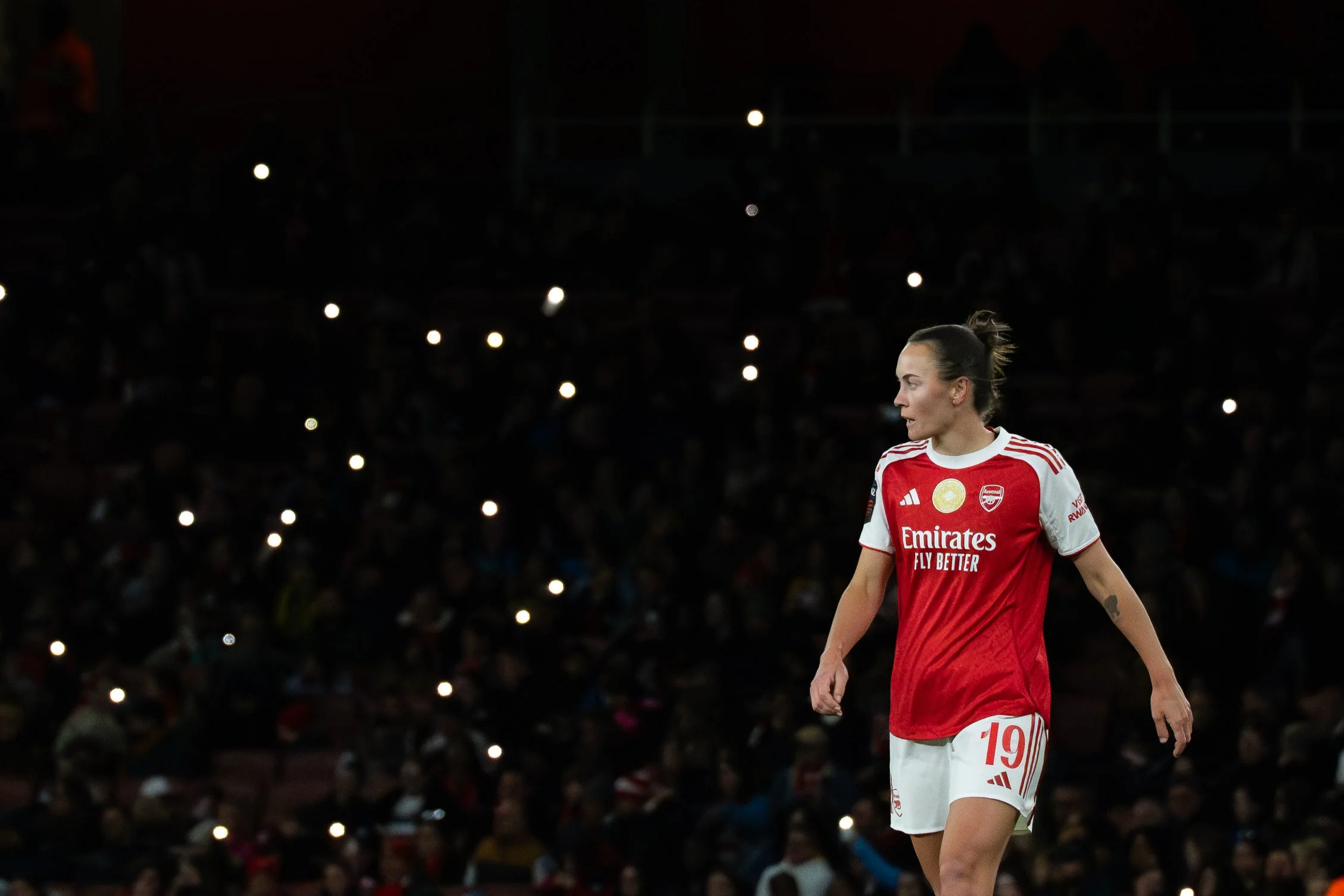 A female soccer player in a red and white uniform walking on the field at night with a crowd and bright lights in the background.