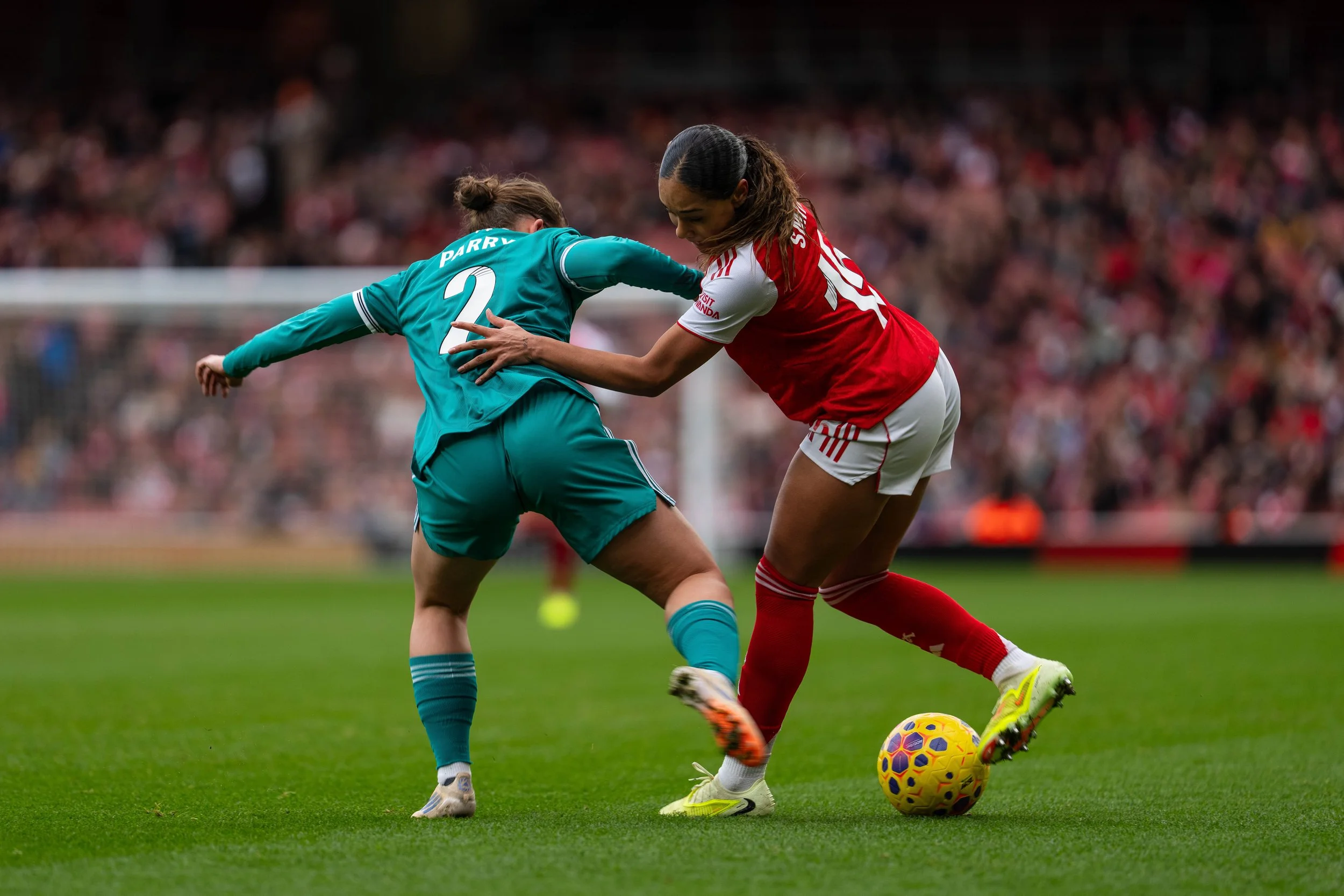 Two female soccer players competing for ball during a match, one in teal jersey and the other in red and white jersey, on a green soccer field with a crowd in the background.