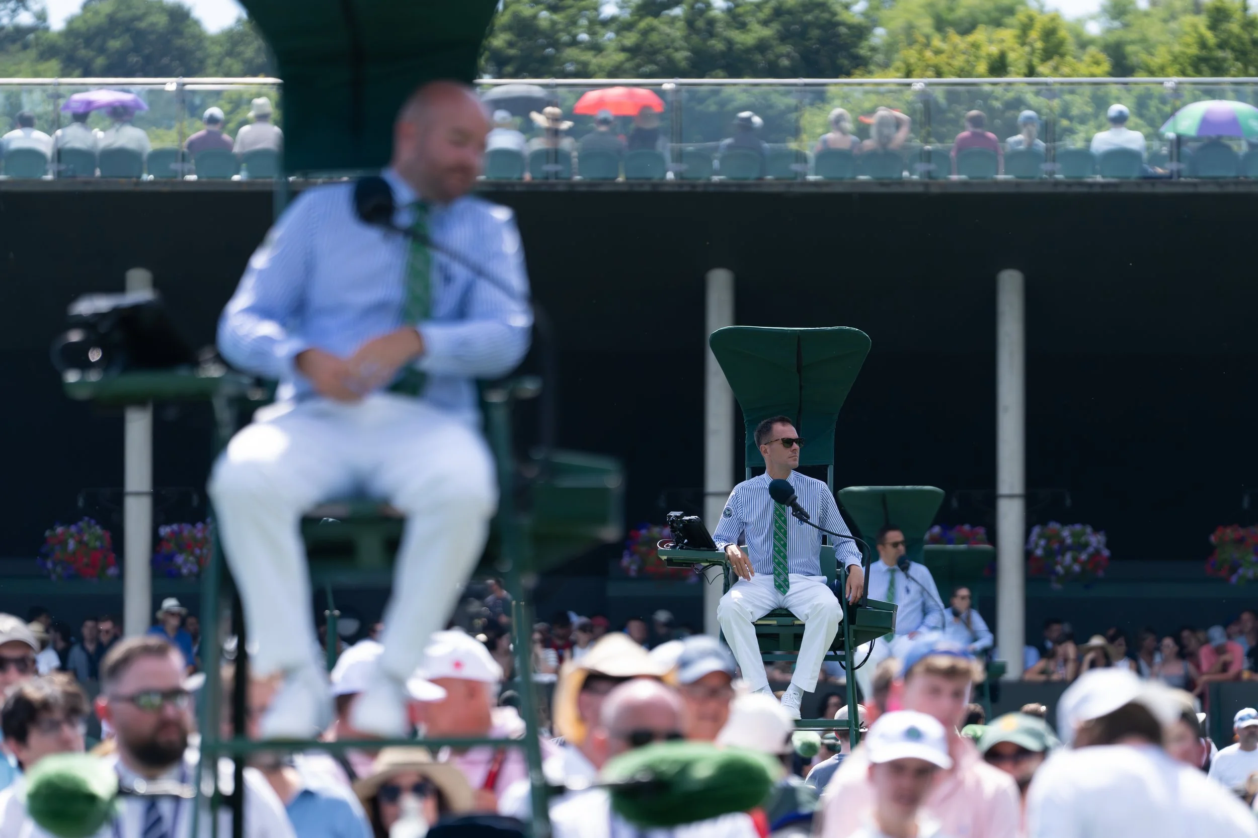 A crowd watching a tennis match at an outdoor stadium with officials sitting on high chairs, some wearing striped shirts and green ties. Spectators are seated behind a glass barrier, with some holding umbrellas, under a bright sunny sky surrounded by green trees.