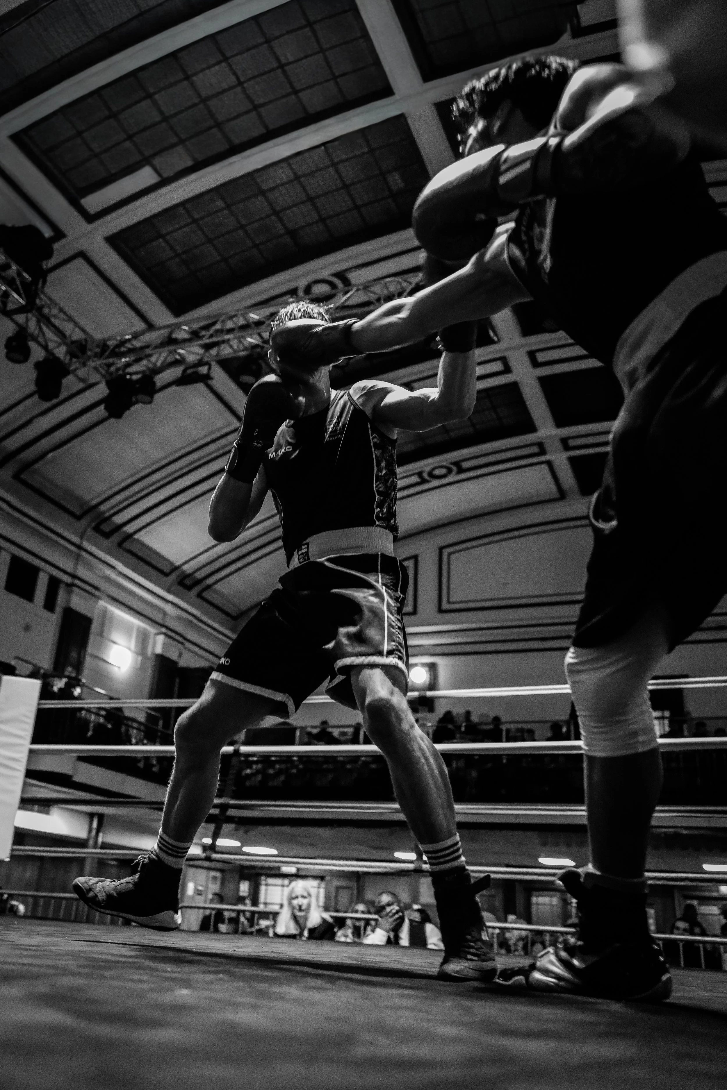 Black and white photo of two amateur boxers in the ring, one delivering a punch to the other. The scene is inside a venue with a high arched ceiling and an audience watching.