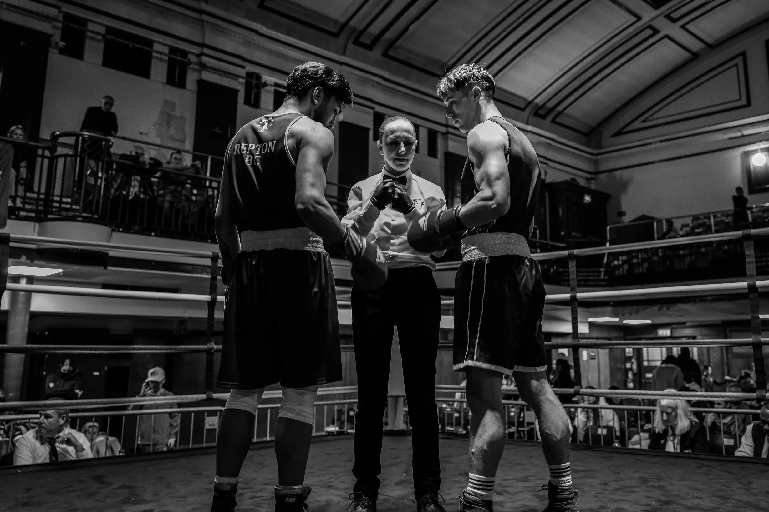Two boxers in black trunks and gloves consulting with a referee in a boxing ring, with spectators watching from the background.