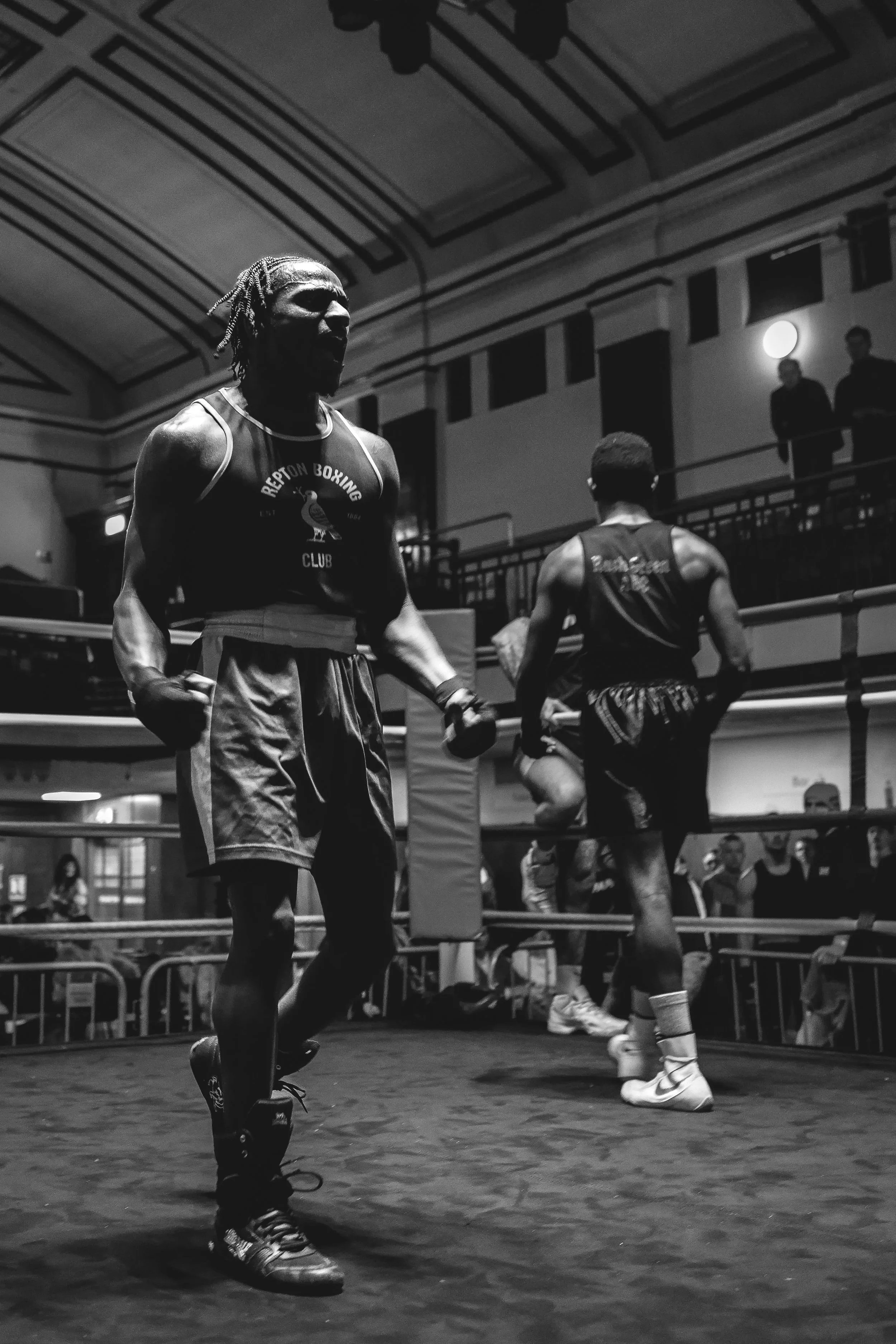 Black and white photo of a boxer in the ring with his fists clenched, wearing sports attire and boxing shoes, with a boxer in the background, and spectators in the stands.