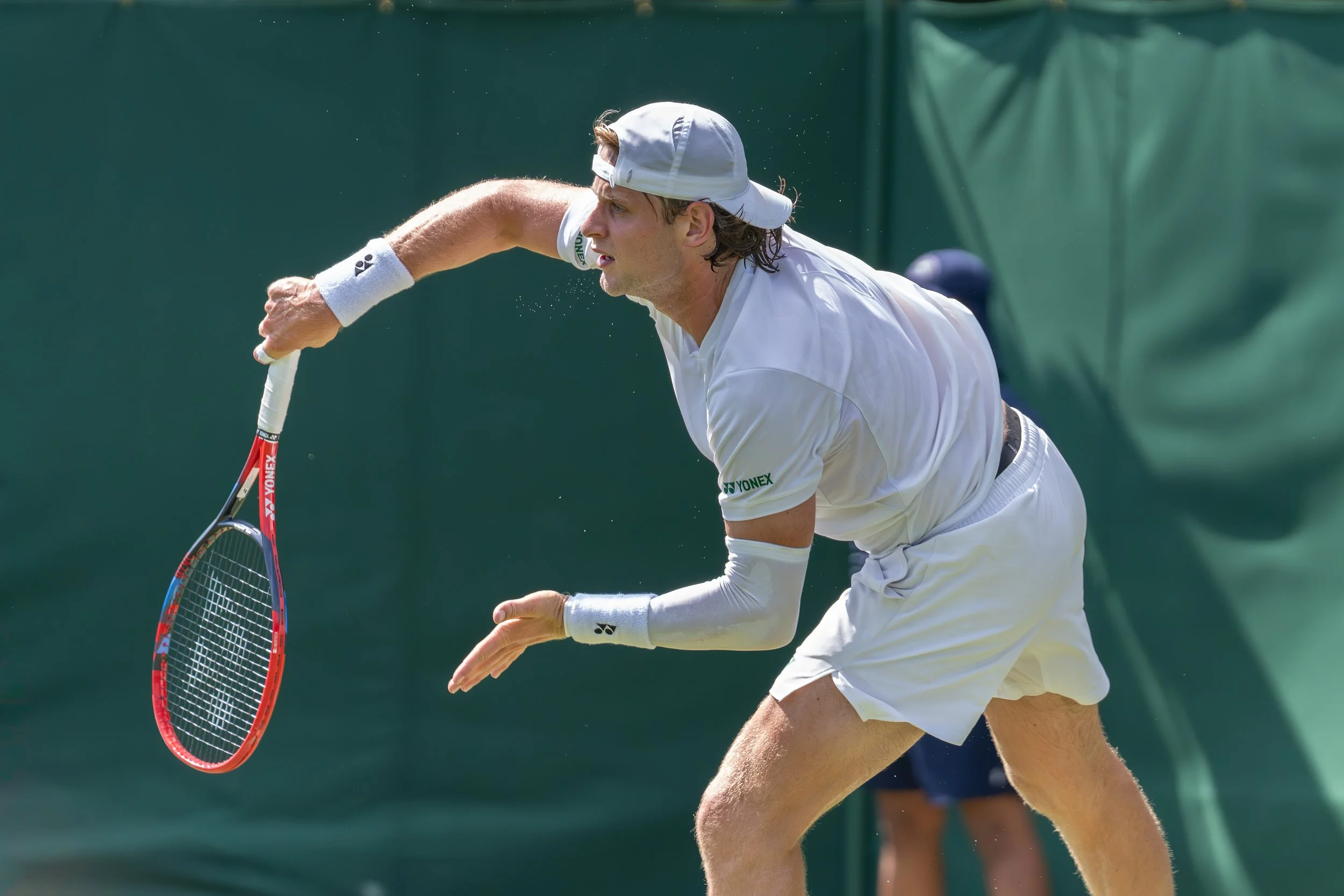 Tennis player in white outfit and cap hitting the ball on court.