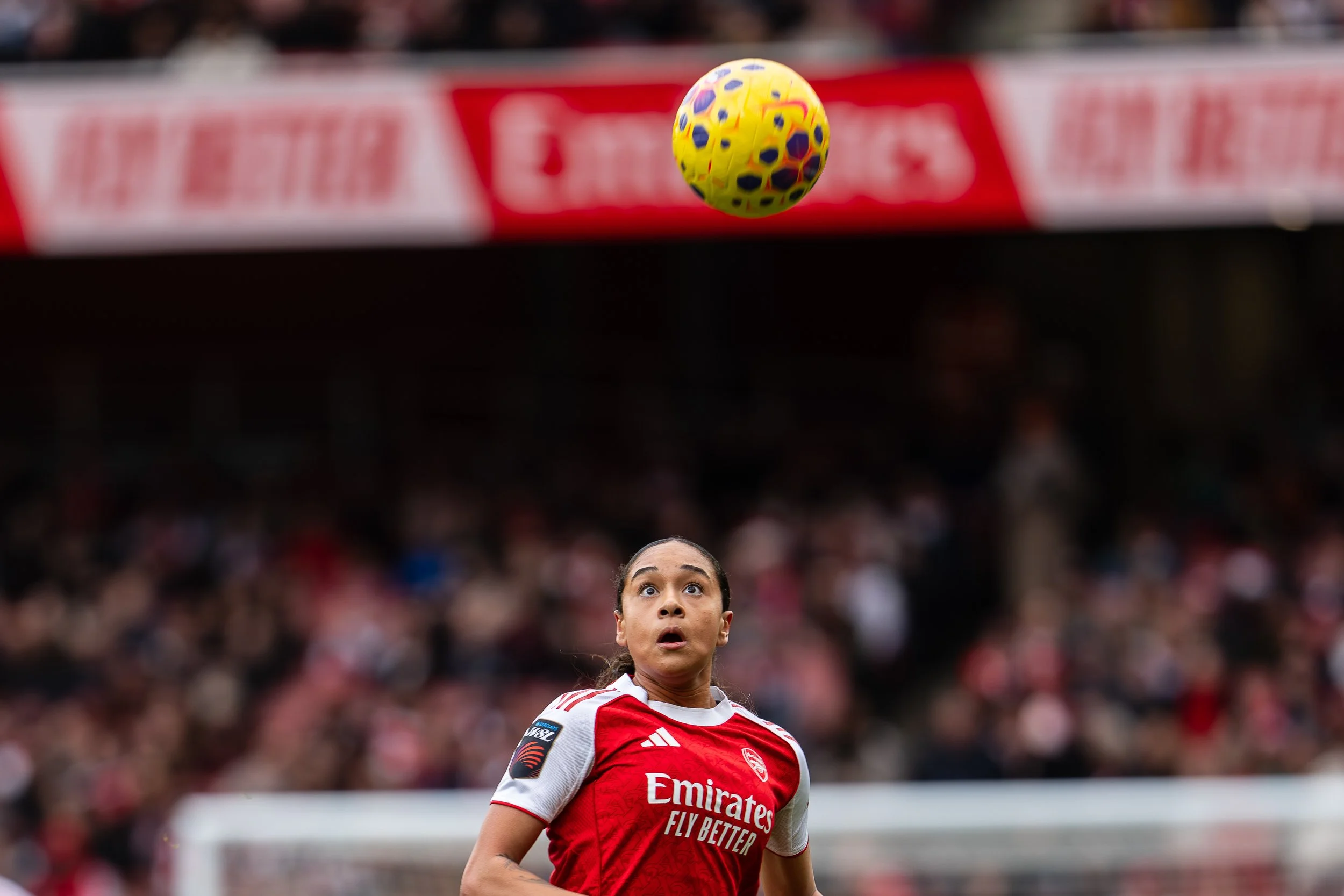 A woman soccer player in a red and white jersey looking up at a yellow and purple soccer ball in a stadium filled with spectators.