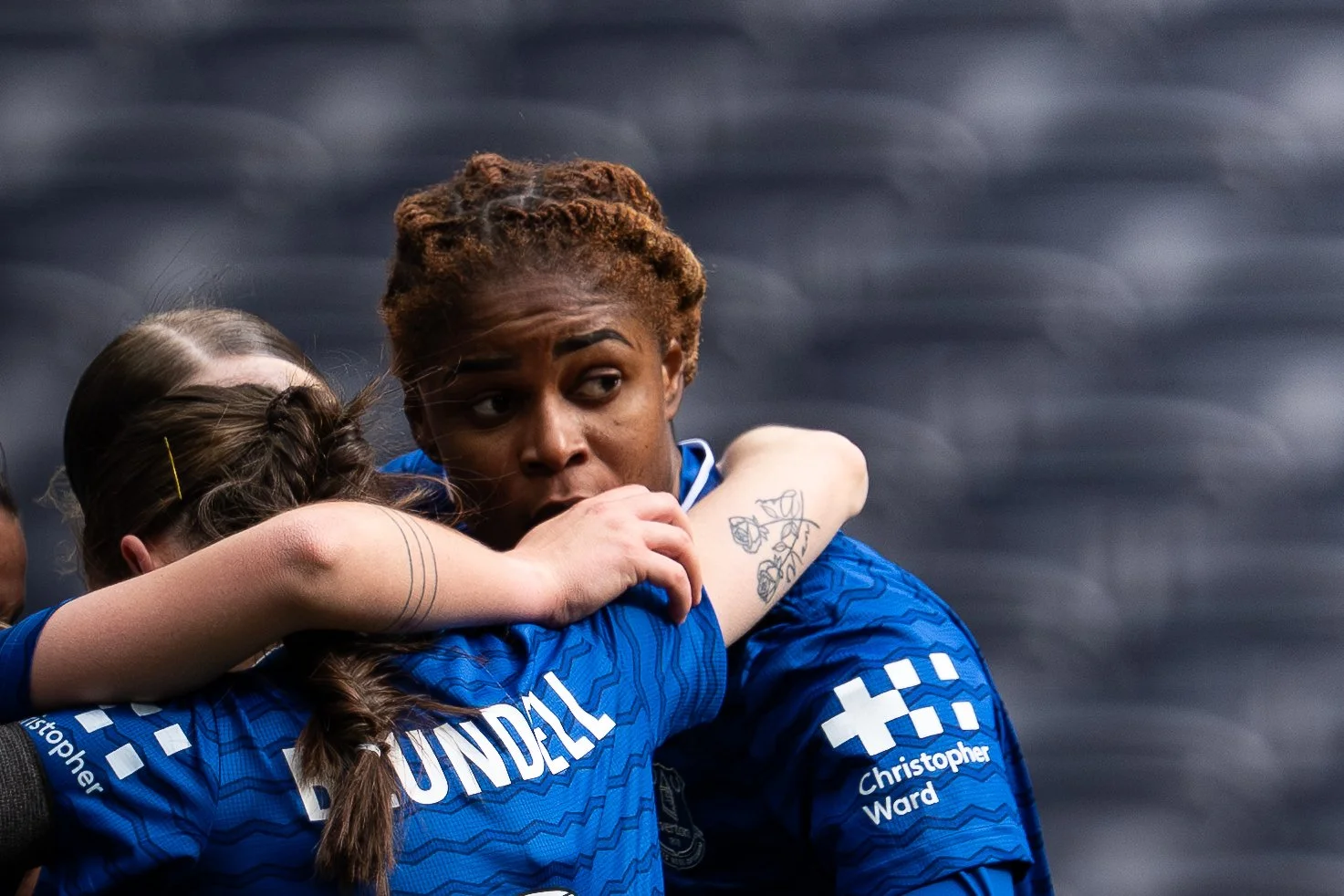 Two female athletes, wearing blue jerseys, embrace each other during a moment of support or celebration, with one showing a focused facial expression.