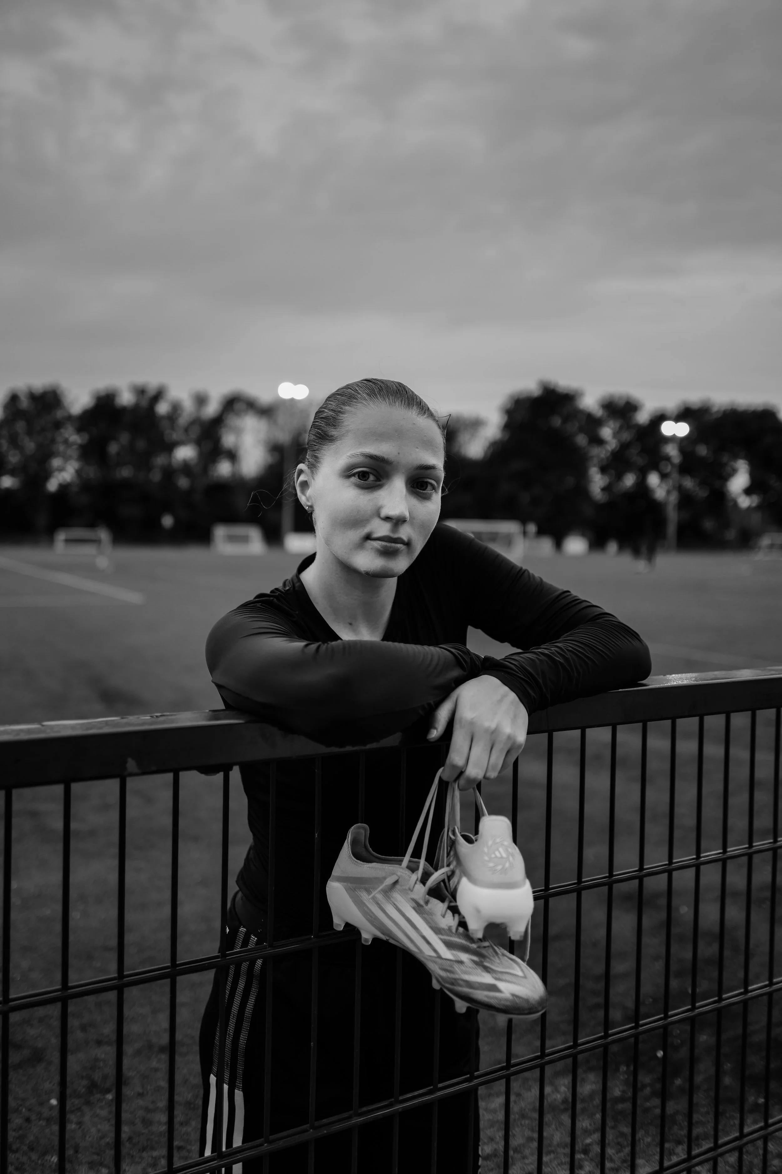 A woman leaning on a fence at an outdoor sports field during dusk, holding a pair of athletic shoes and a water bottle, with cloudy skies and trees in the background.