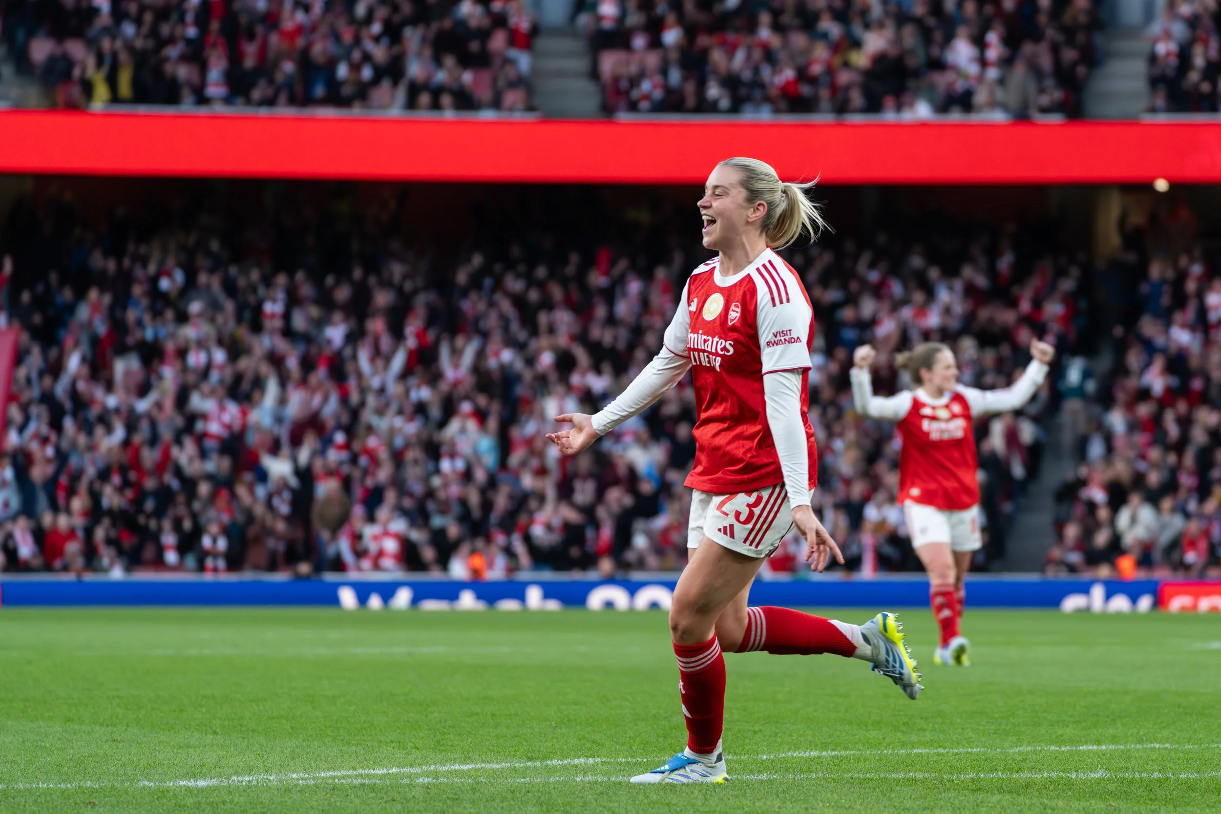 A female soccer player wearing a red and white uniform celebrating on a soccer field with a large crowd in the background.