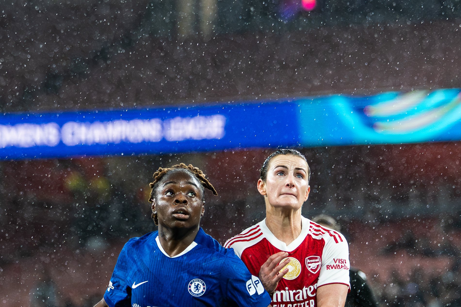 Two female soccer players competing in a match under a rainy sky; one player is wearing a blue jersey with the Chelsea football club logo, and the other is wearing a red and white jersey with the Arsenal football club logo. Both players are looking upward, focused, with rain falling around them.