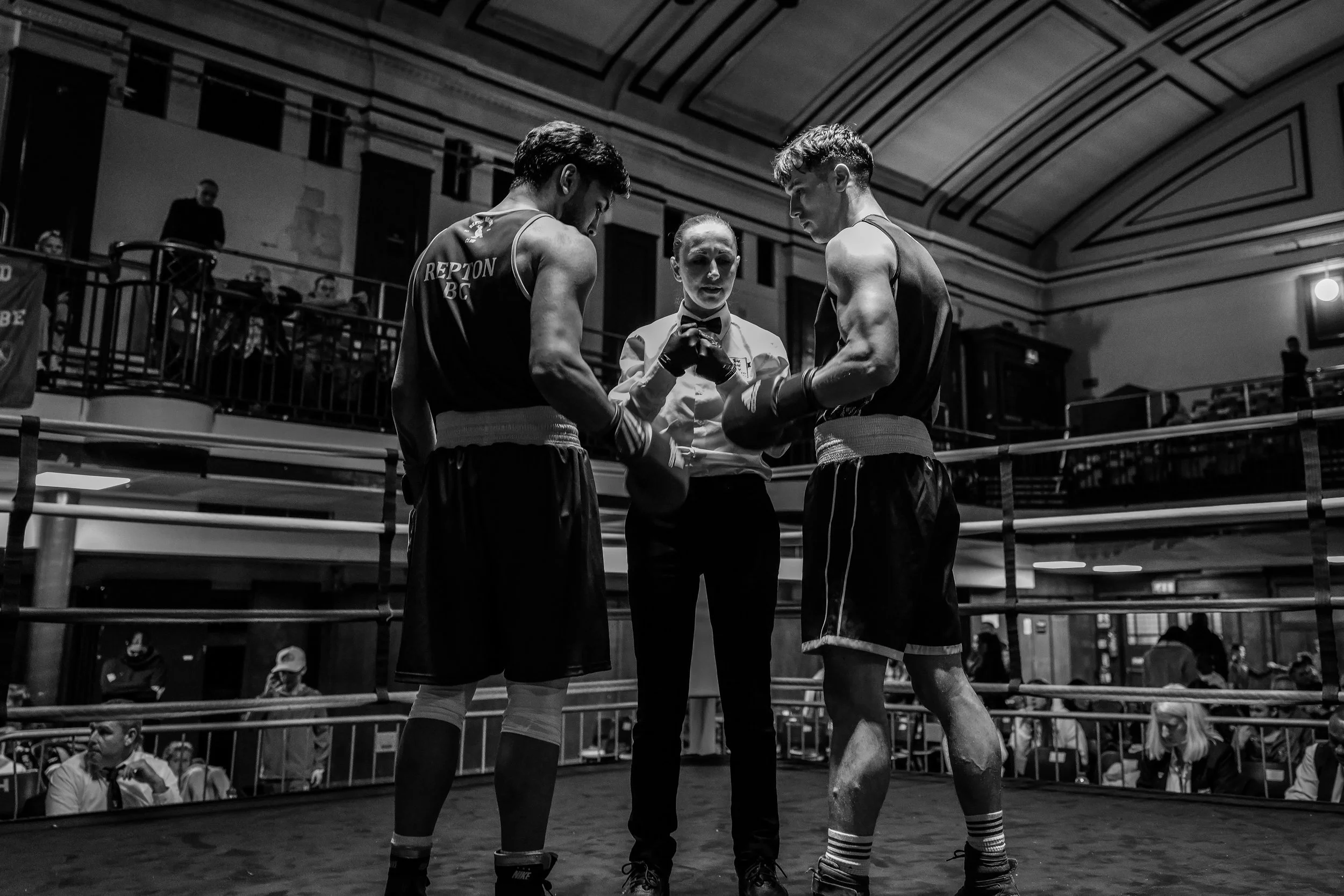 Two male boxers in a ring, standing face to face, with a female referee between them, inspecting or listening to their instructions in a boxing match. The scene is in a boxing venue with spectators seated around.