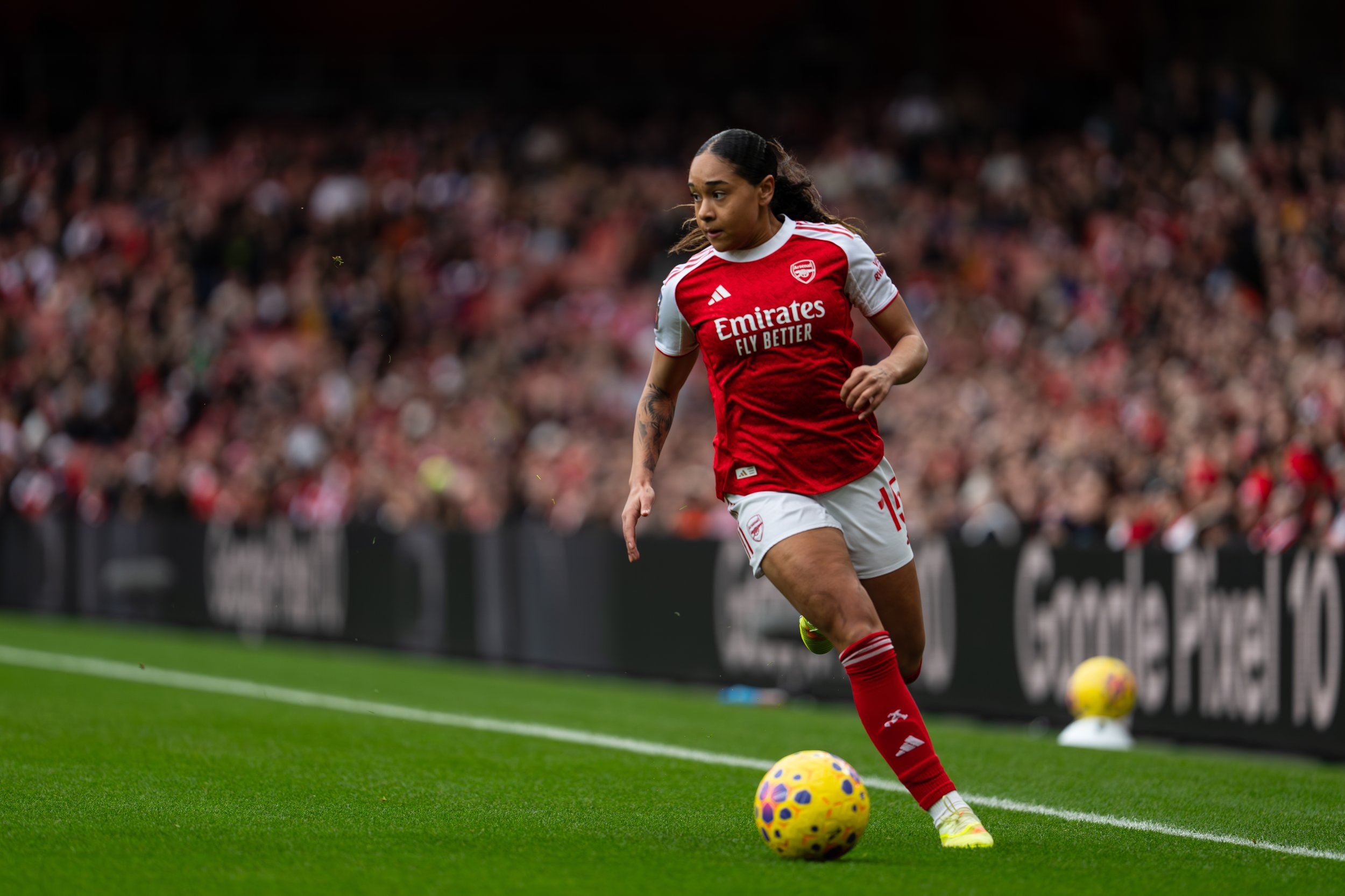 Female soccer player in red and white uniform preparing to kick a yellow and purple soccer ball during a match, with a blurred crowd in the background.