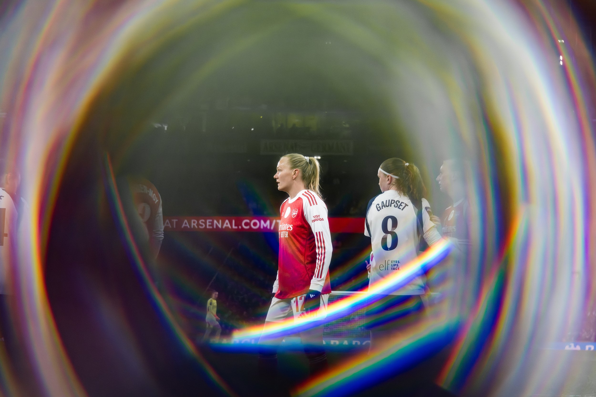 View through a colorful, circular object showing female soccer players, one in a red jersey and another in a white jersey, on a soccer field with an advertisement banner in the background.
