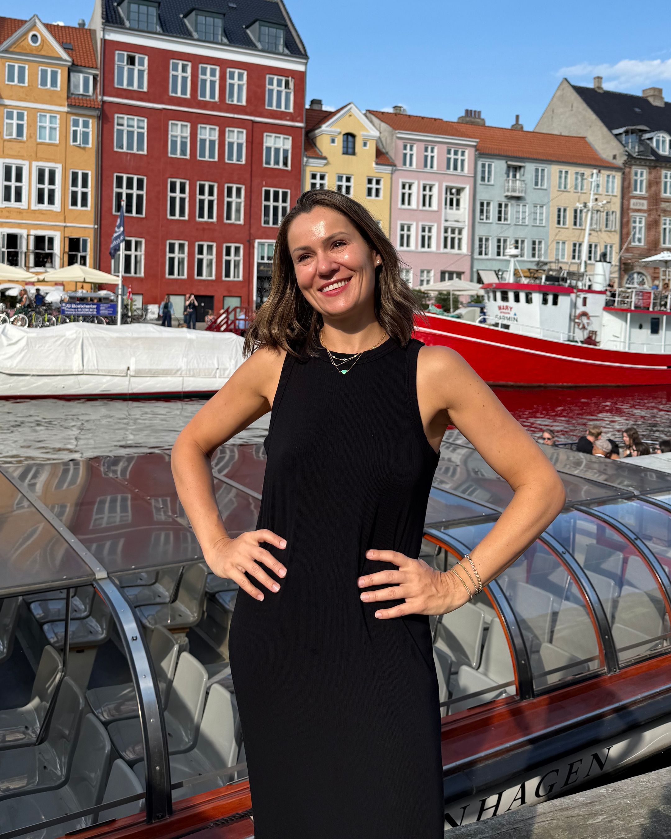 Marina Cooley smiling in Copenhagen, Denmark. She is wearing a black sleeveless dress and standing in front of colorful canal houses and a red boat.