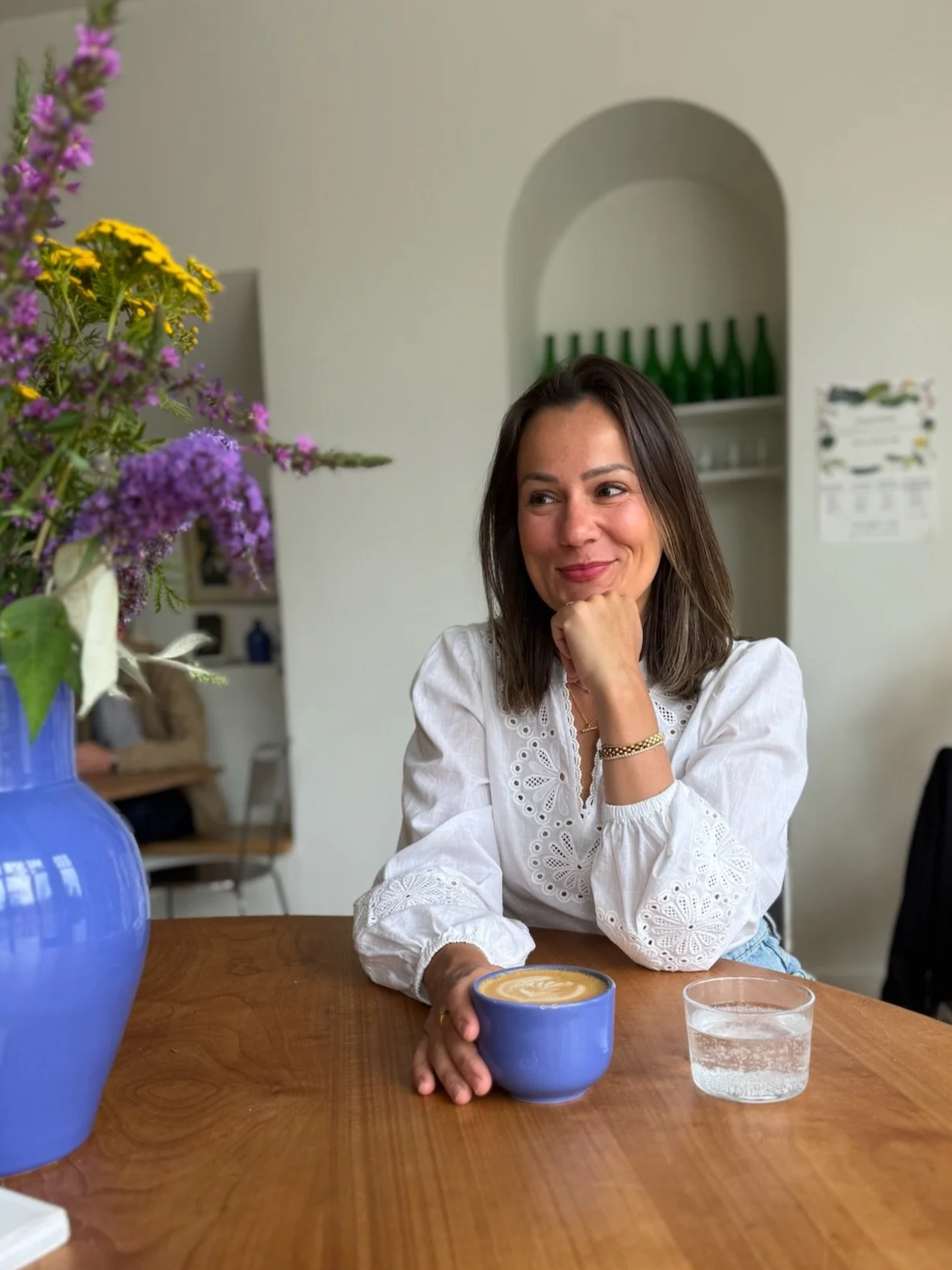 Marina Cooley sits at a wooden table with a blue cup of coffee and a large blue vase with purple and yellow flowers. She is smiling and resting her chin on her hand. 