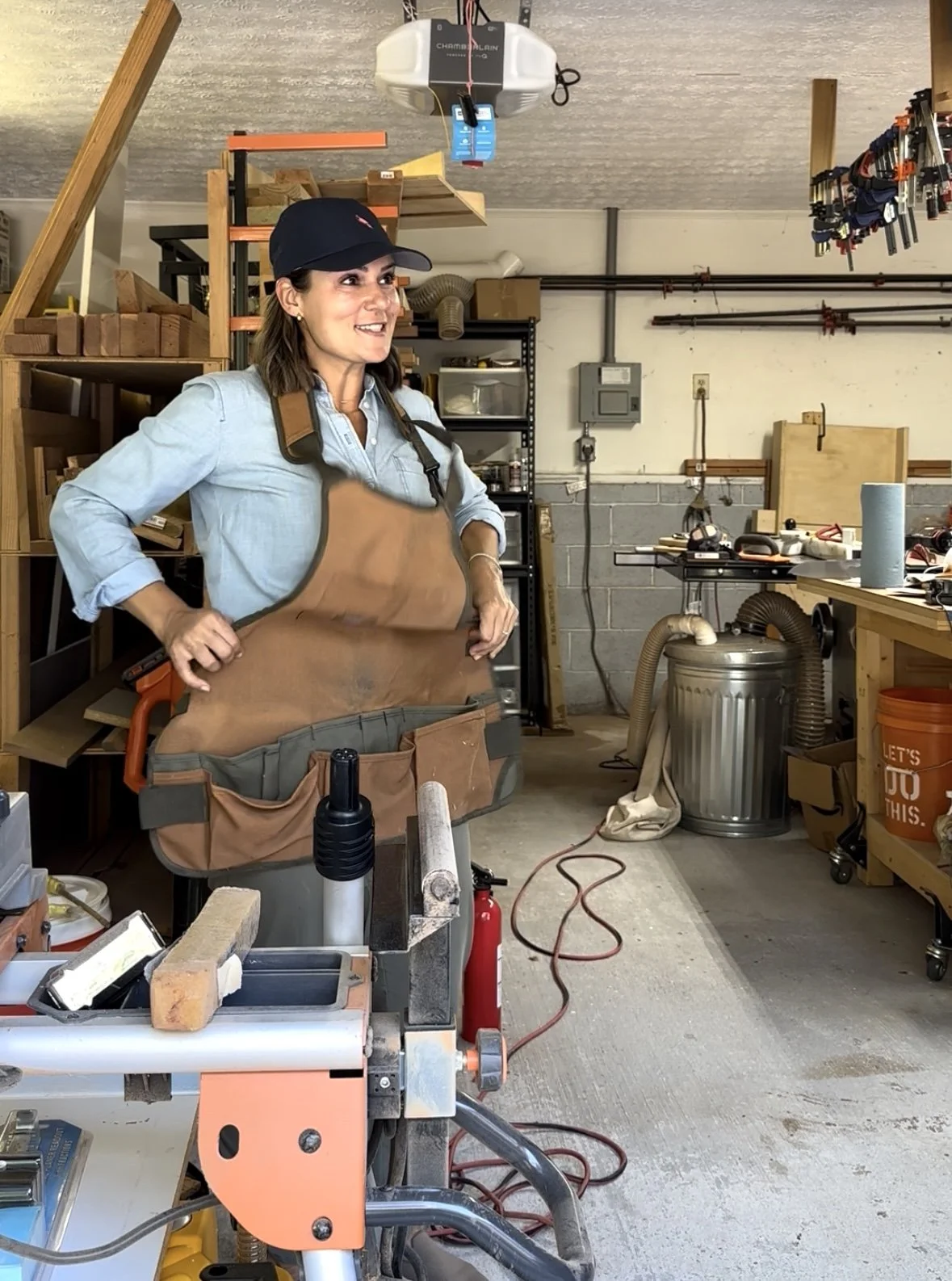 Marina Cooley in a workshop wearing a cap and apron, standing near a workbench with tools, woodworking equipment, and storage shelves in the background.