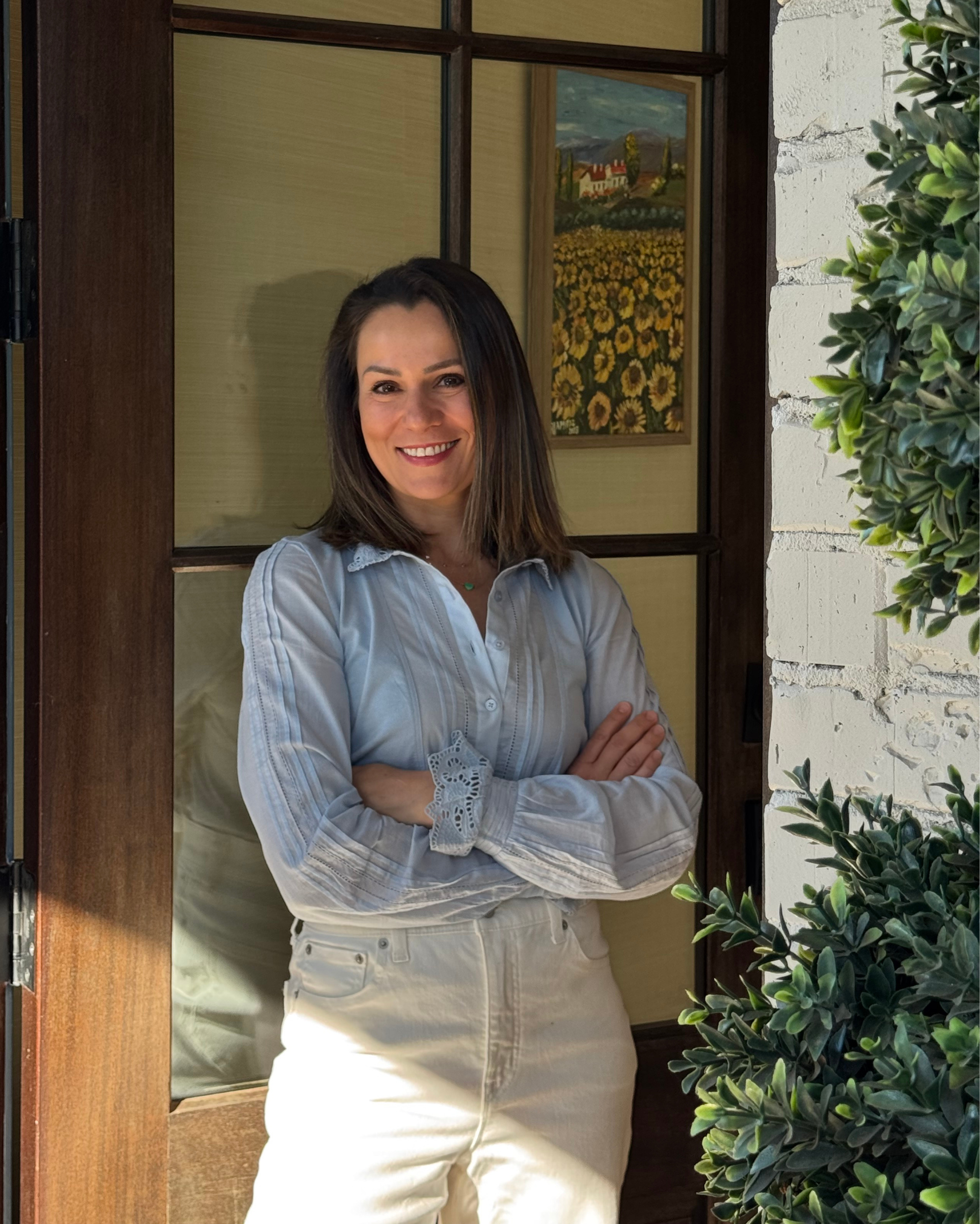 Marina Cooley standing outside, smiling with arms crossed, wearing a light-colored blouse and light pants, next to a white brick wall and lush green bushes, with a doorway in the background.